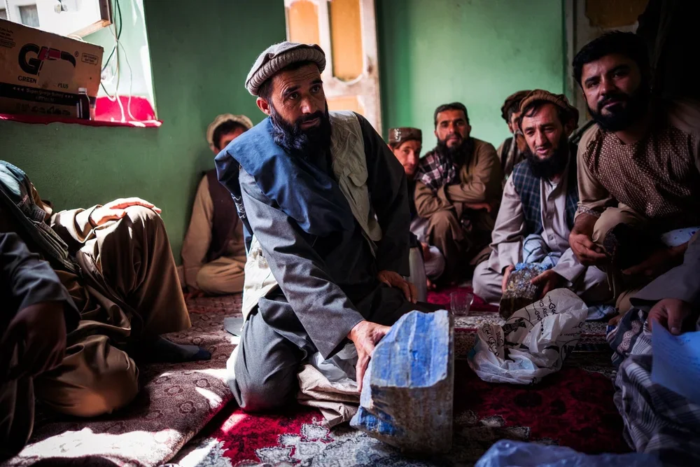 A group of men gathered closely inside a room with green walls, sitting on the floor on a patterned carpet. One man in the center is kneeling and holding a large rock of lapis lazuli, while the others watch and listen attentively.