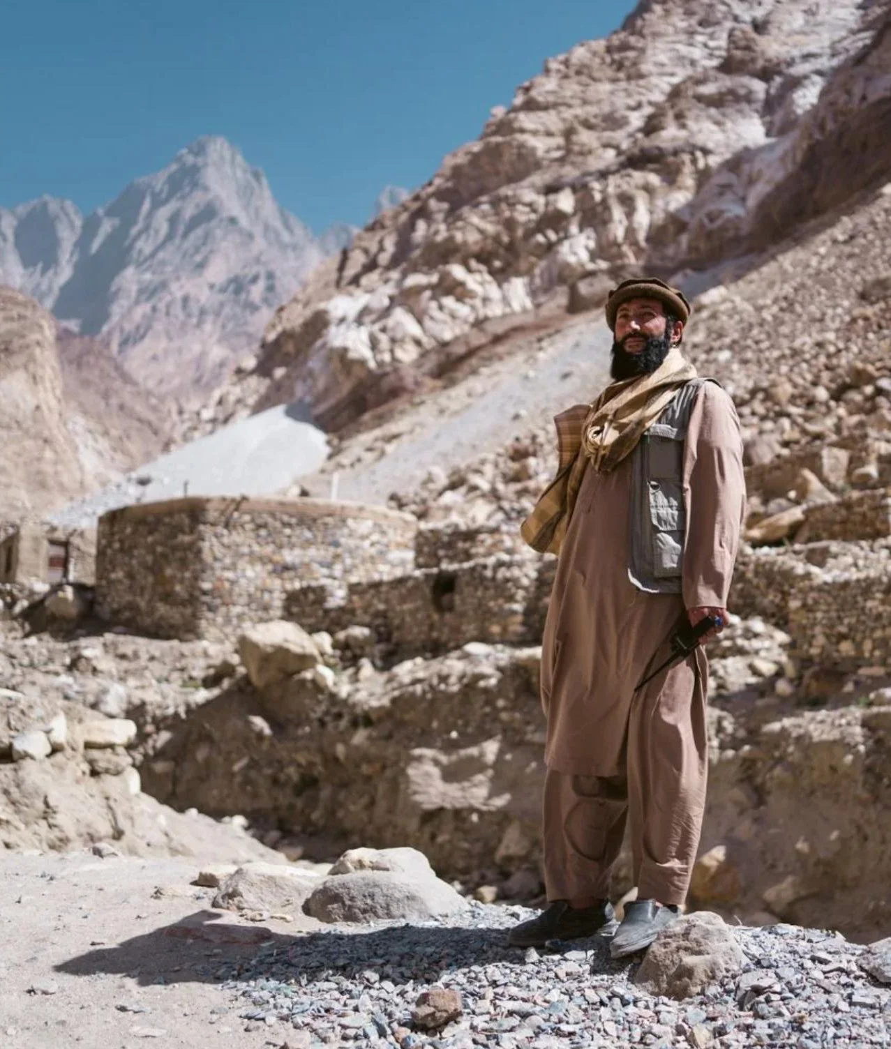 A man dressed in traditional Middle Eastern clothing standing in a mountainous desert landscape with rocky cliffs and snow-capped peaks in the background.