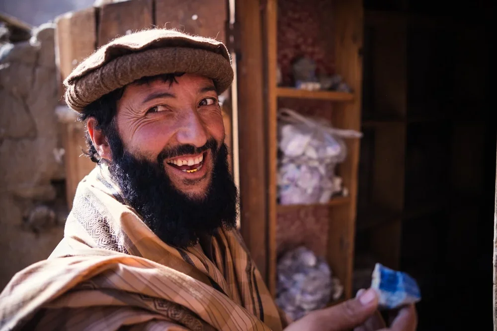 A man with a beard, wearing a hat and traditional clothing, smiling and holding a piece of lapis inside a rustic room with wooden shelves and stone walls.
