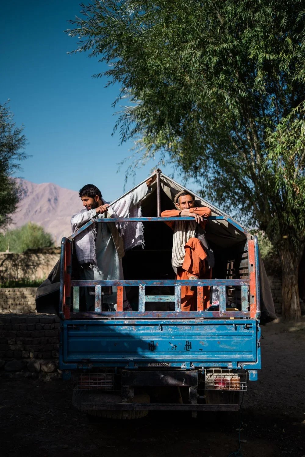 Two men standing in the back of a blue truck, with one leaning on the side and smiling, and the other resting his arms on the top of the truck, under a large leafy tree with mountains in the background.