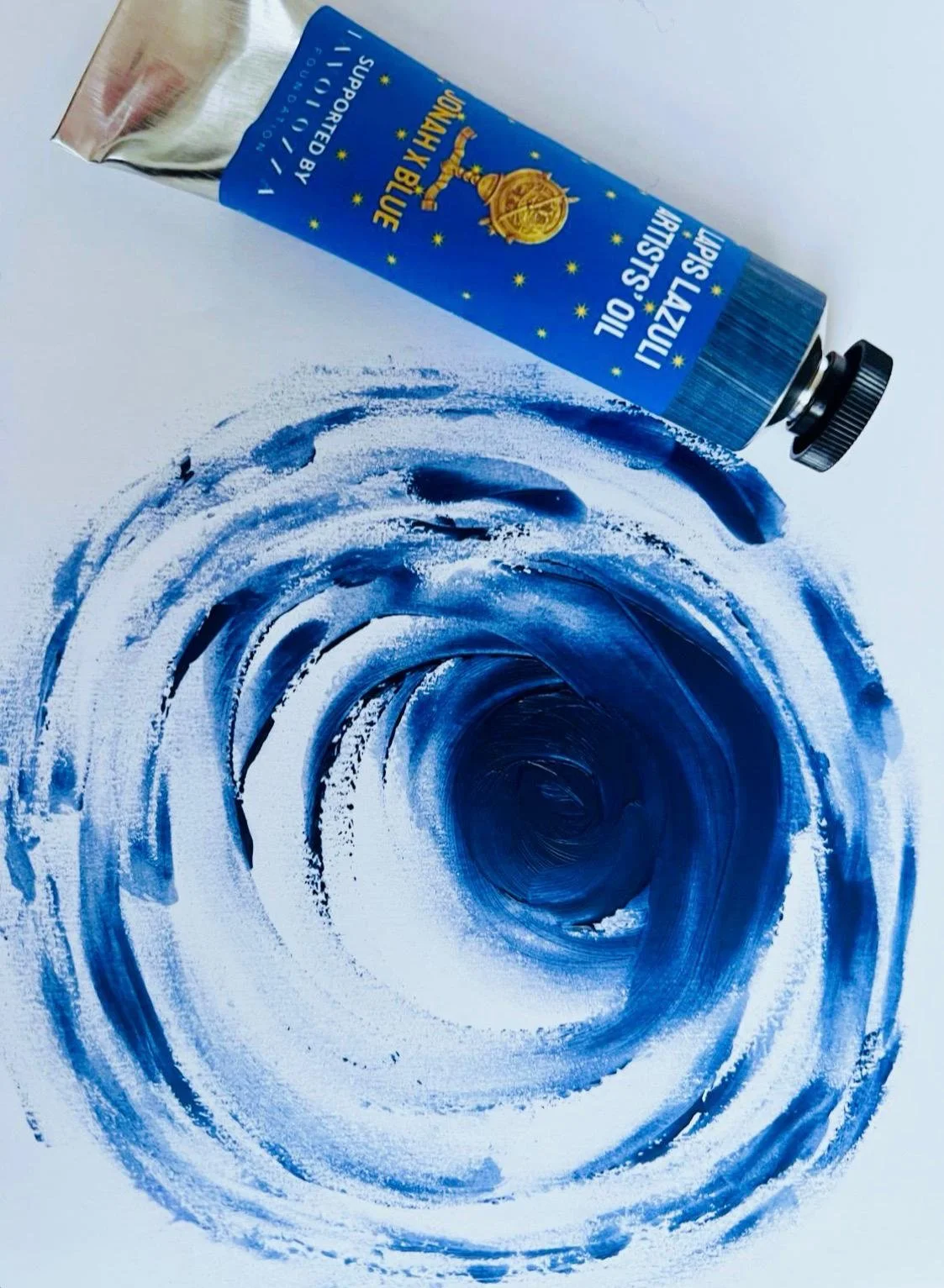 A tube of Jonah x Blue oil paint by Cranfield Colours resting horizontally on a white surface, with a circular swirl of blue oil paint on the surface beneath it.