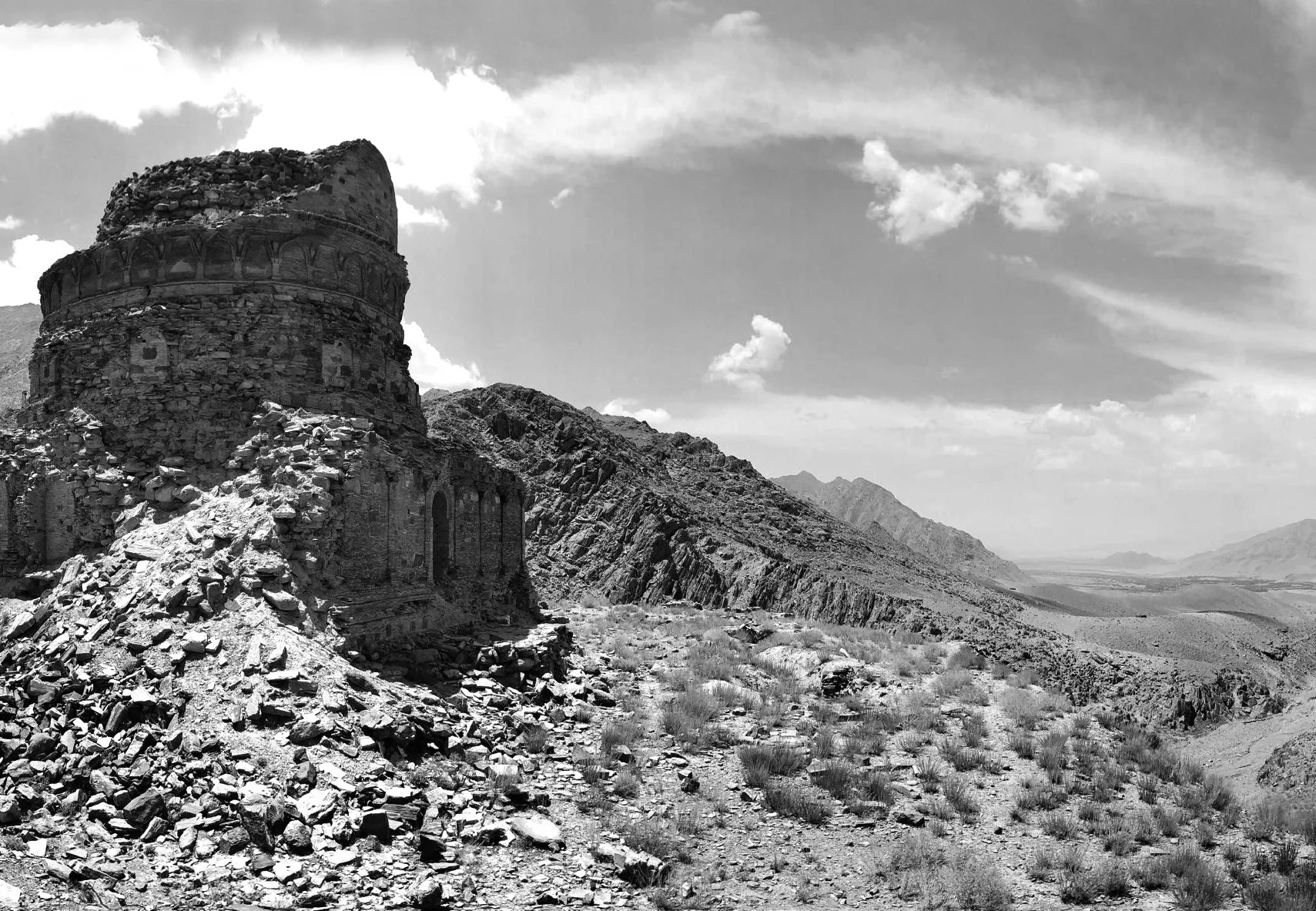 Black and white photograph of an ancient, partially ruined stone structure on a rocky, barren hillside with distant mountains and a partly cloudy sky in the background.