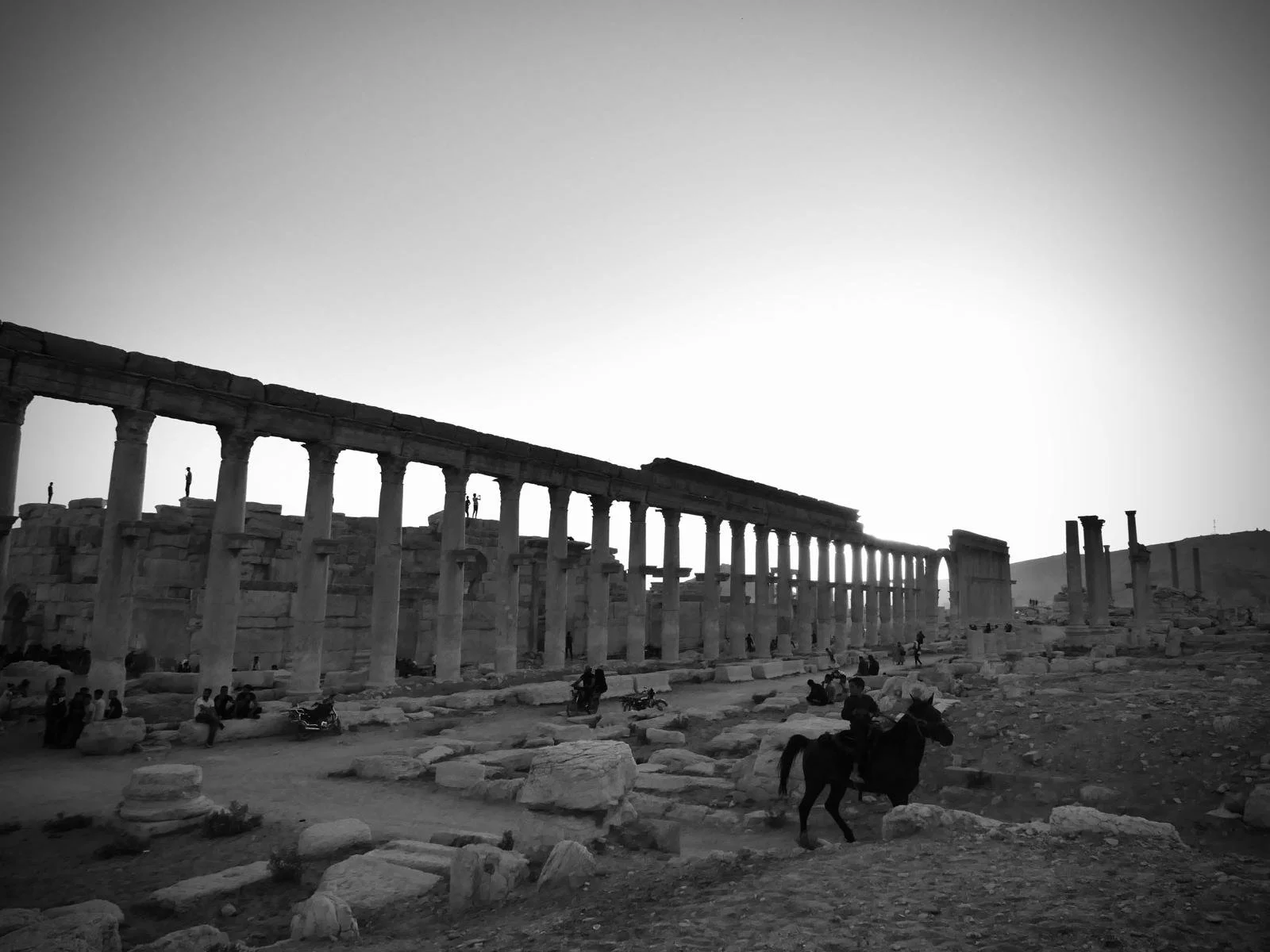 Black and white photo of ancient ruins with tall columns, people walking and standing around, and a person riding a horse in the foreground