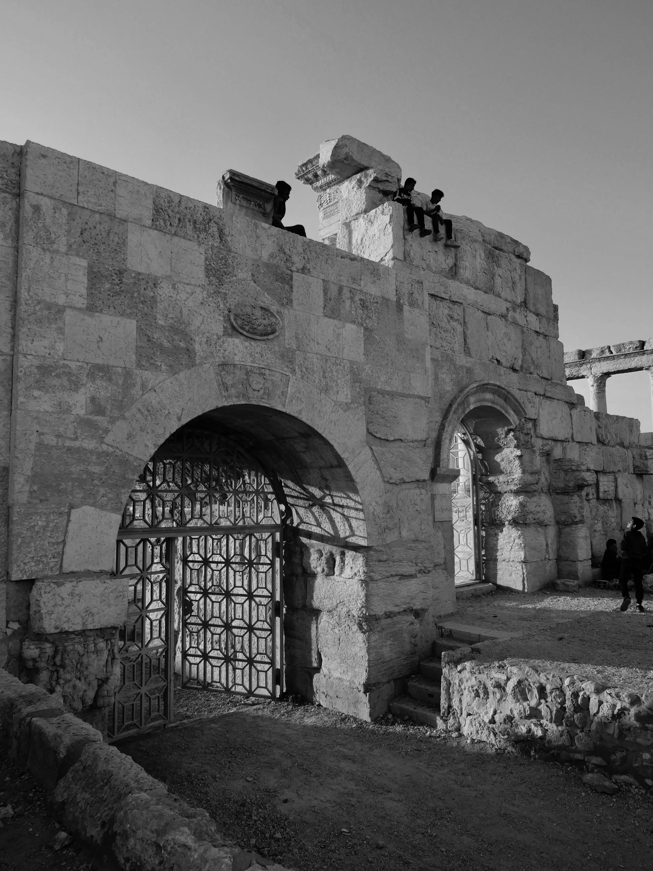 Black and white photo of an ancient stone structure with arched doorways, metal gates, and children sitting on the stone remains at the top of the structure.