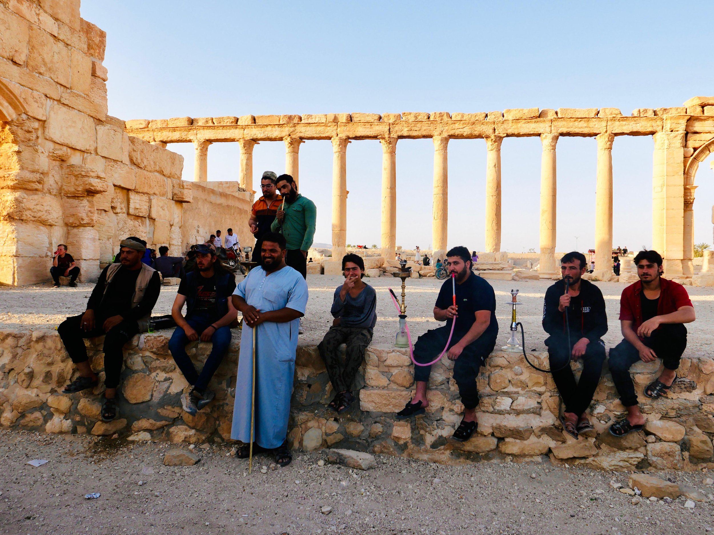 Group of people sitting and standing on a stone wall in front of ancient ruins with tall columns, people in the background, under a clear sky.