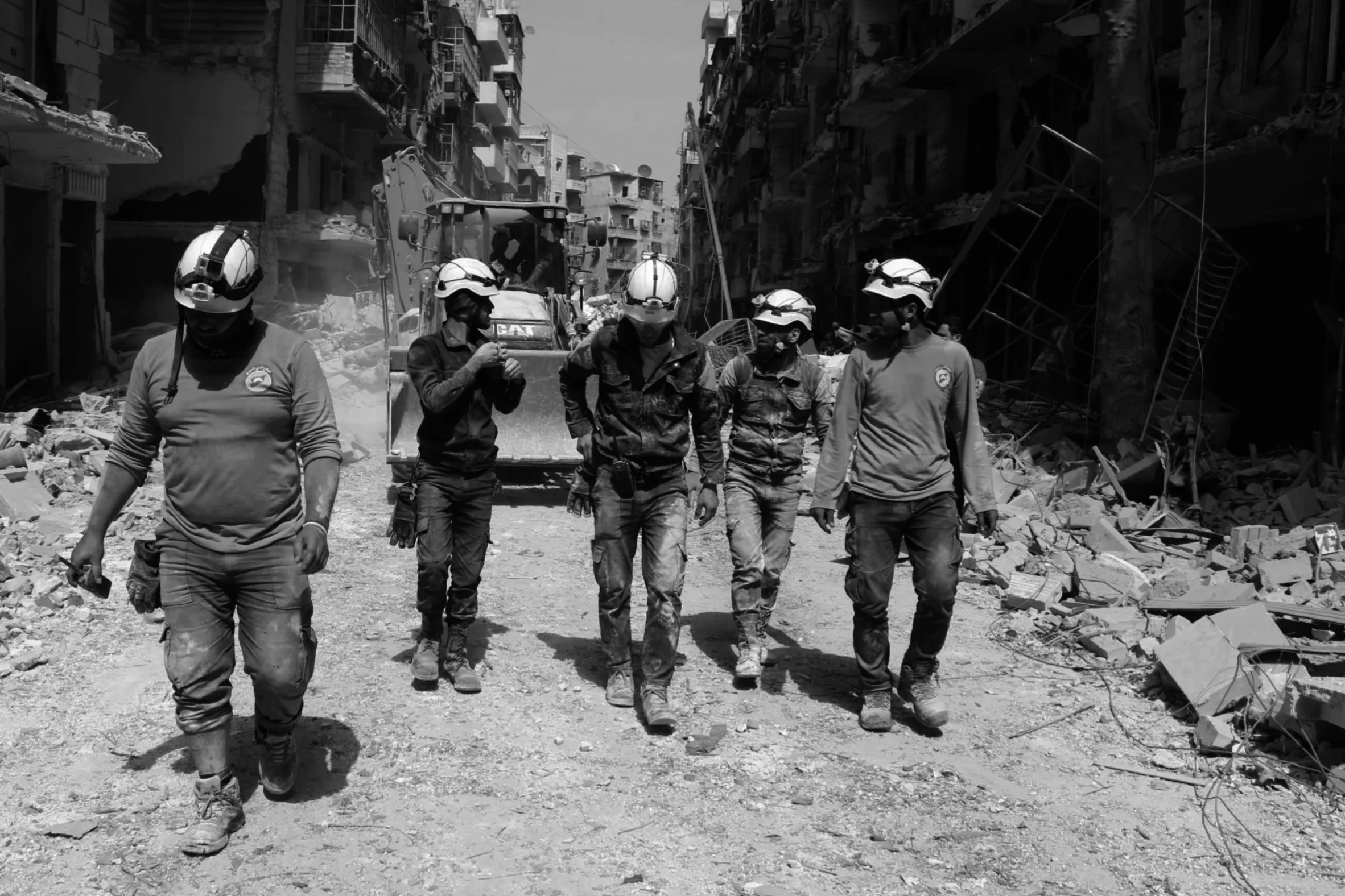 Six White Helmets walking through a rubble-strewn street with damaged buildings and construction equipment in the background.