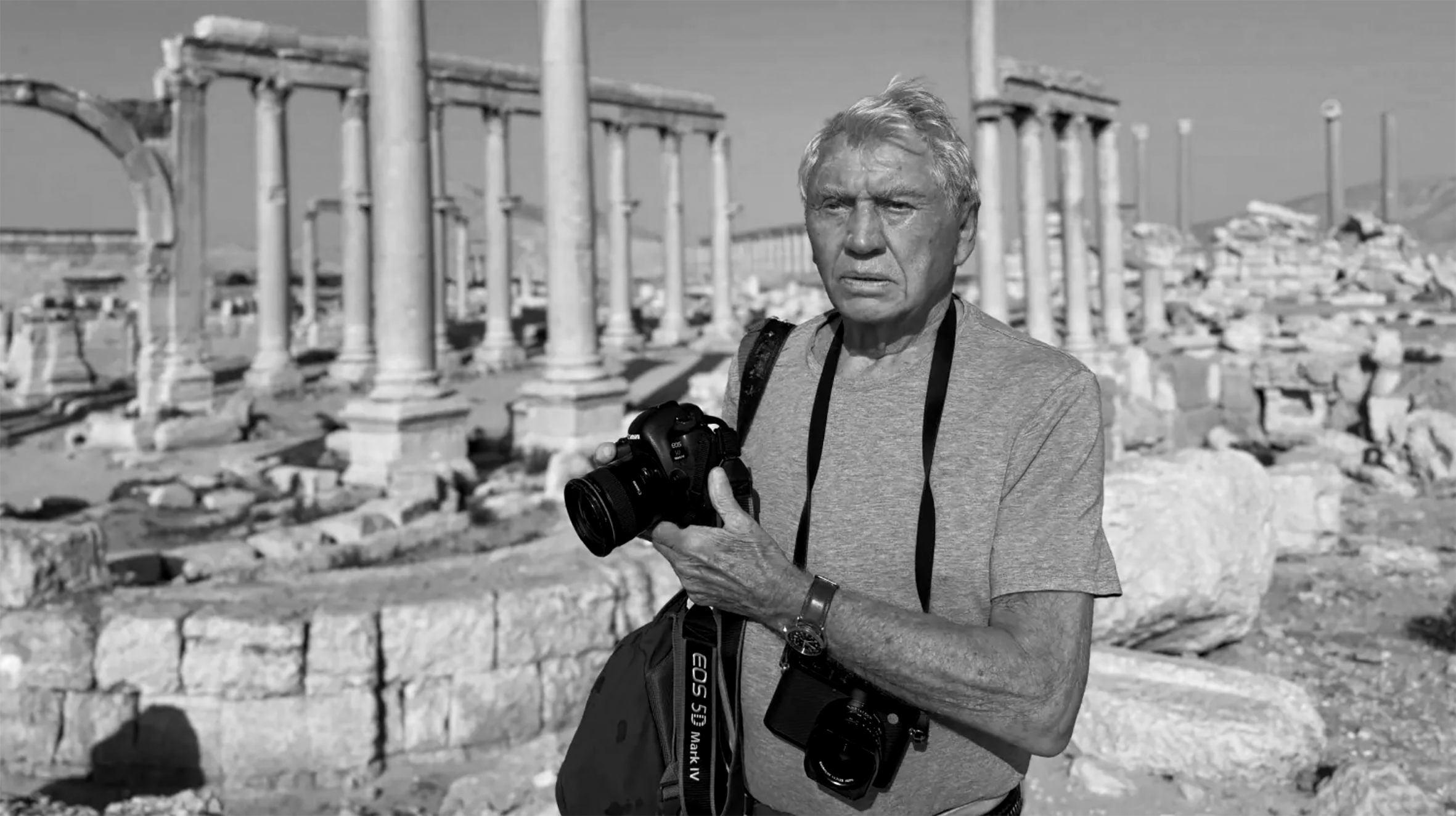 An elderly man with a camera around his neck holding a camera, standing among ancient ruins with columns and stone debris in the background.
