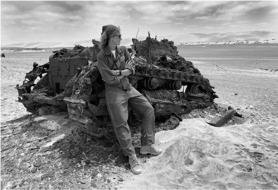Bella Pollen leaning against a wrecked, burned-out tank on a deserted beach with dunes and cloudy sky in the background.