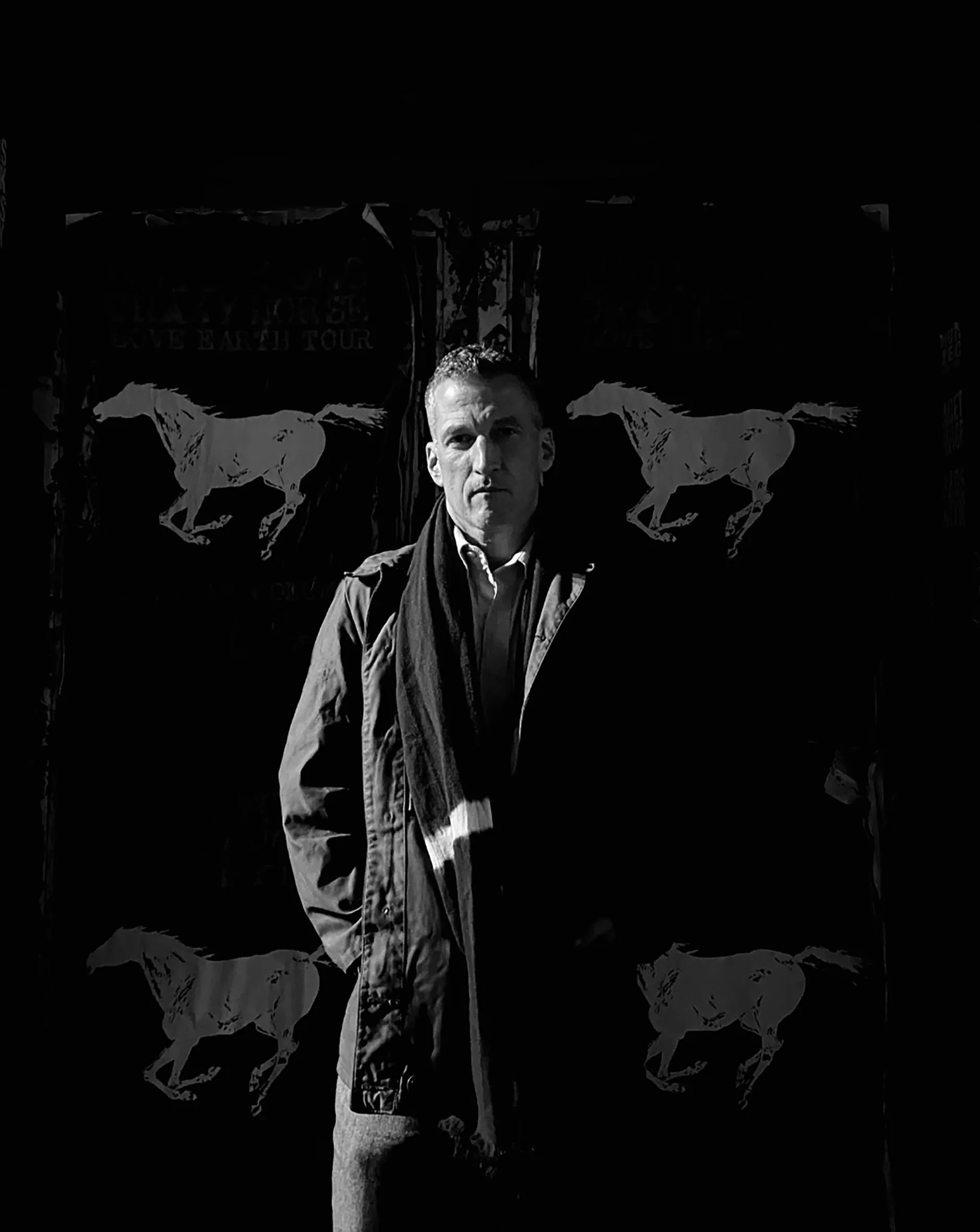 Ben Timberlake stands in front of a background with horse images, wearing a jacket and scarf, with shadowed lighting.