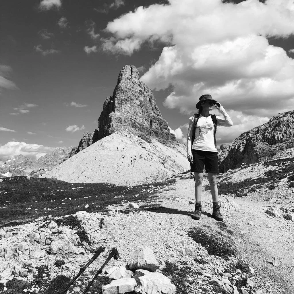 A woman standing on a rocky trail in a mountainous landscape, wearing hiking gear including a hat, sunglasses, and a backpack, with a tall rock formation and clouds in the sky behind her.