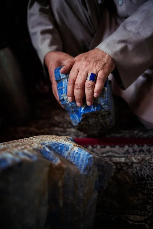 Close-up of a person's hand wearing a blue ring, holding a chisel and carving a blue stone block.