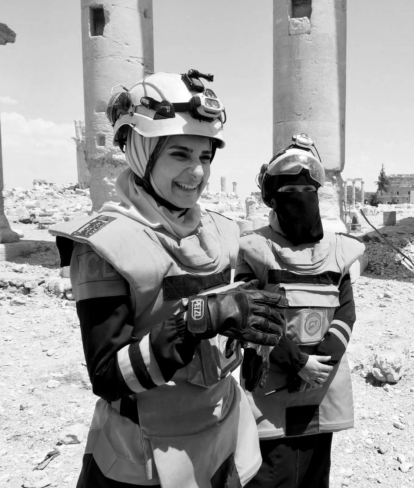 Two rescue workers in protective gear, helmets, and gloves standing amidst ruins of a destroyed building, smiling and appearing to communicate.