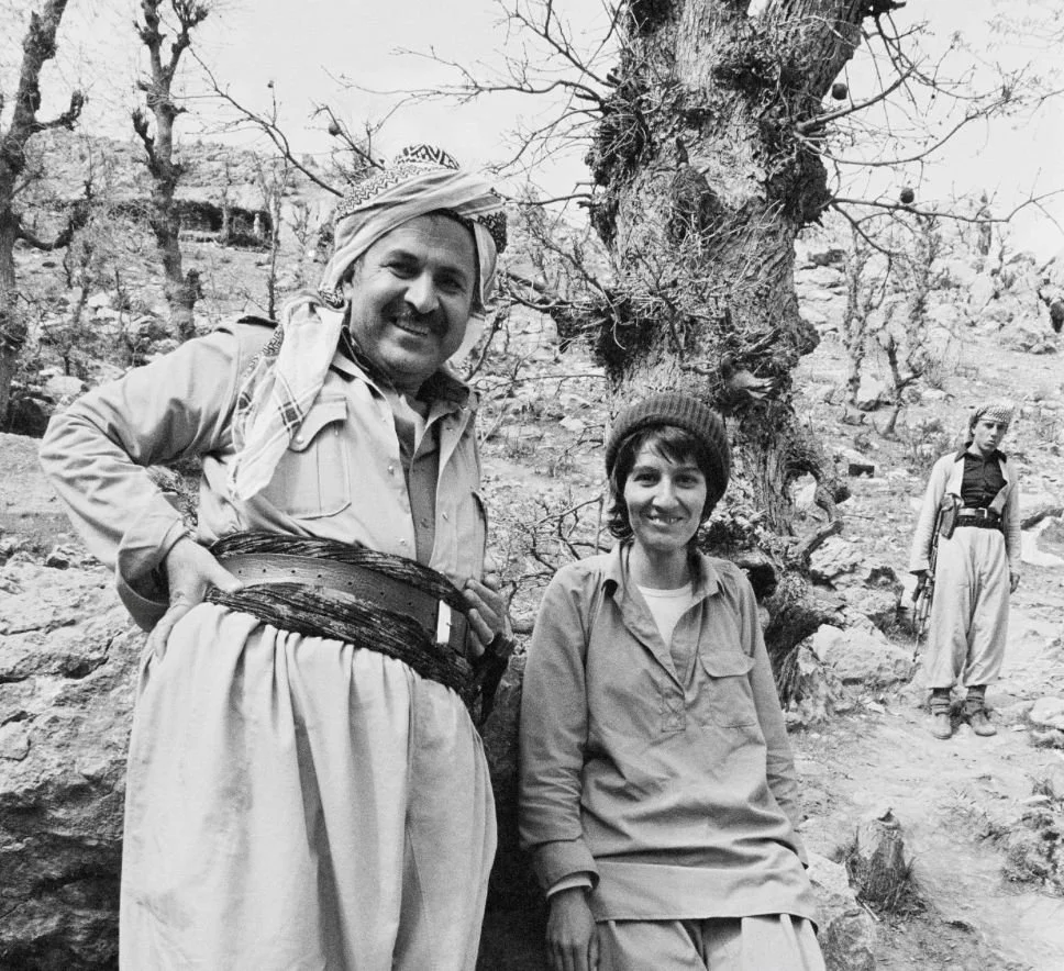 Three people outdoors in a rocky area near a leafless tree. A man with a mustache and a woman with short hair and a beanie sit and stand near the tree, smiling. A woman in the background wears a jacket and pants and looks to the side.