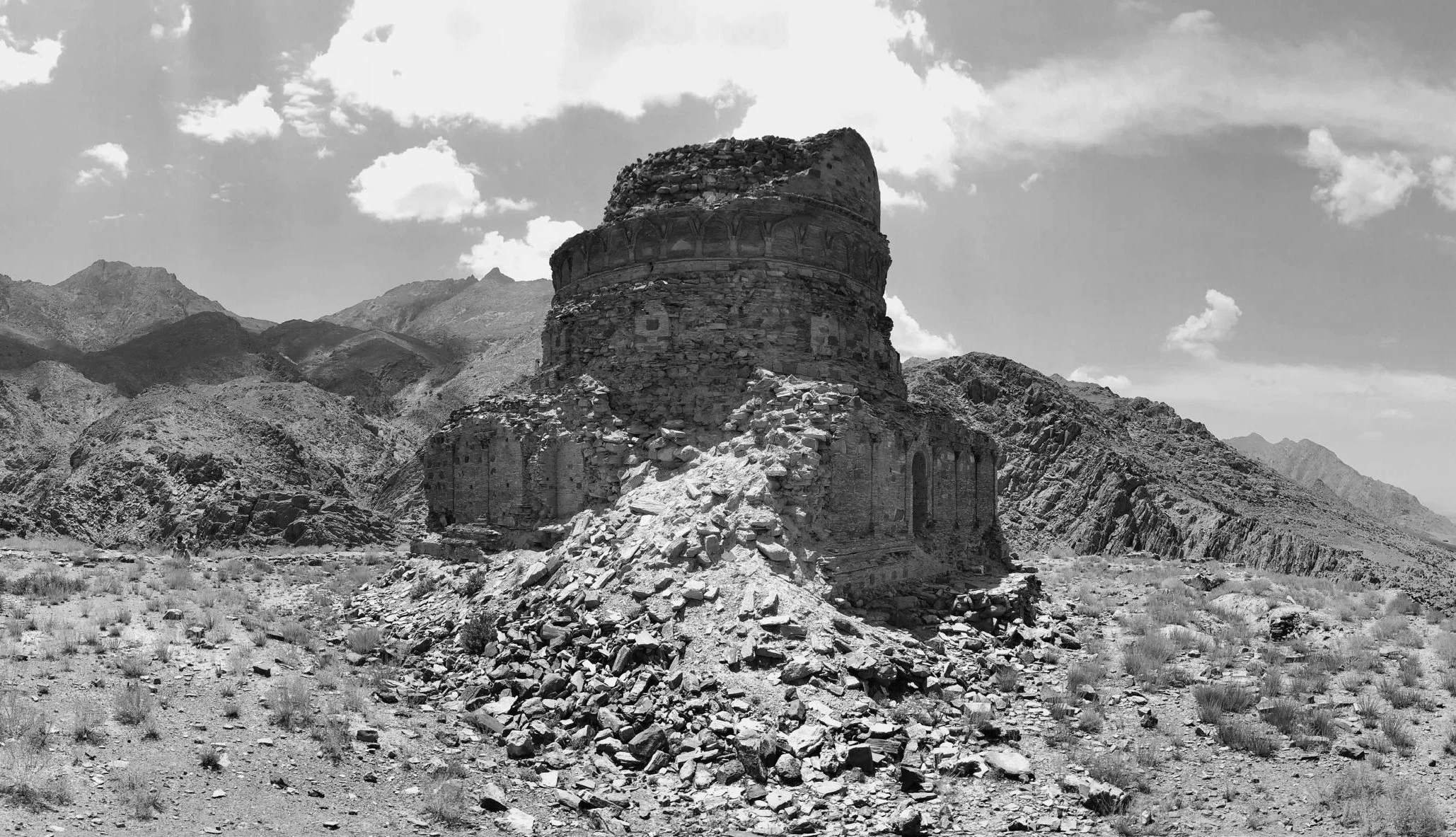 Ruined ancient structure on a rocky plain with mountains in the background, under a partly cloudy sky.