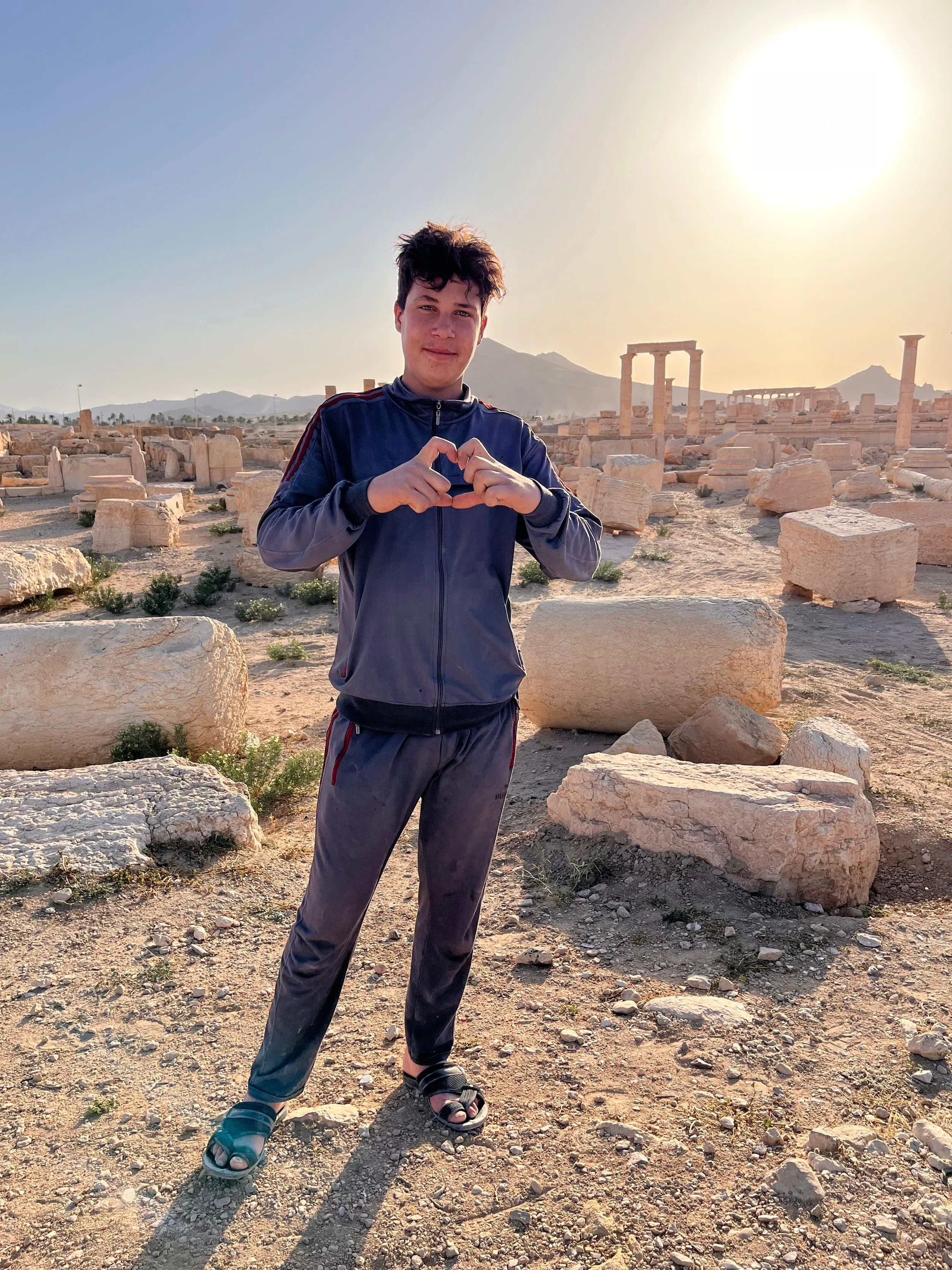 A young man in a gray and black tracksuit making a heart shape with his hands at an ancient ruin site during sunset.