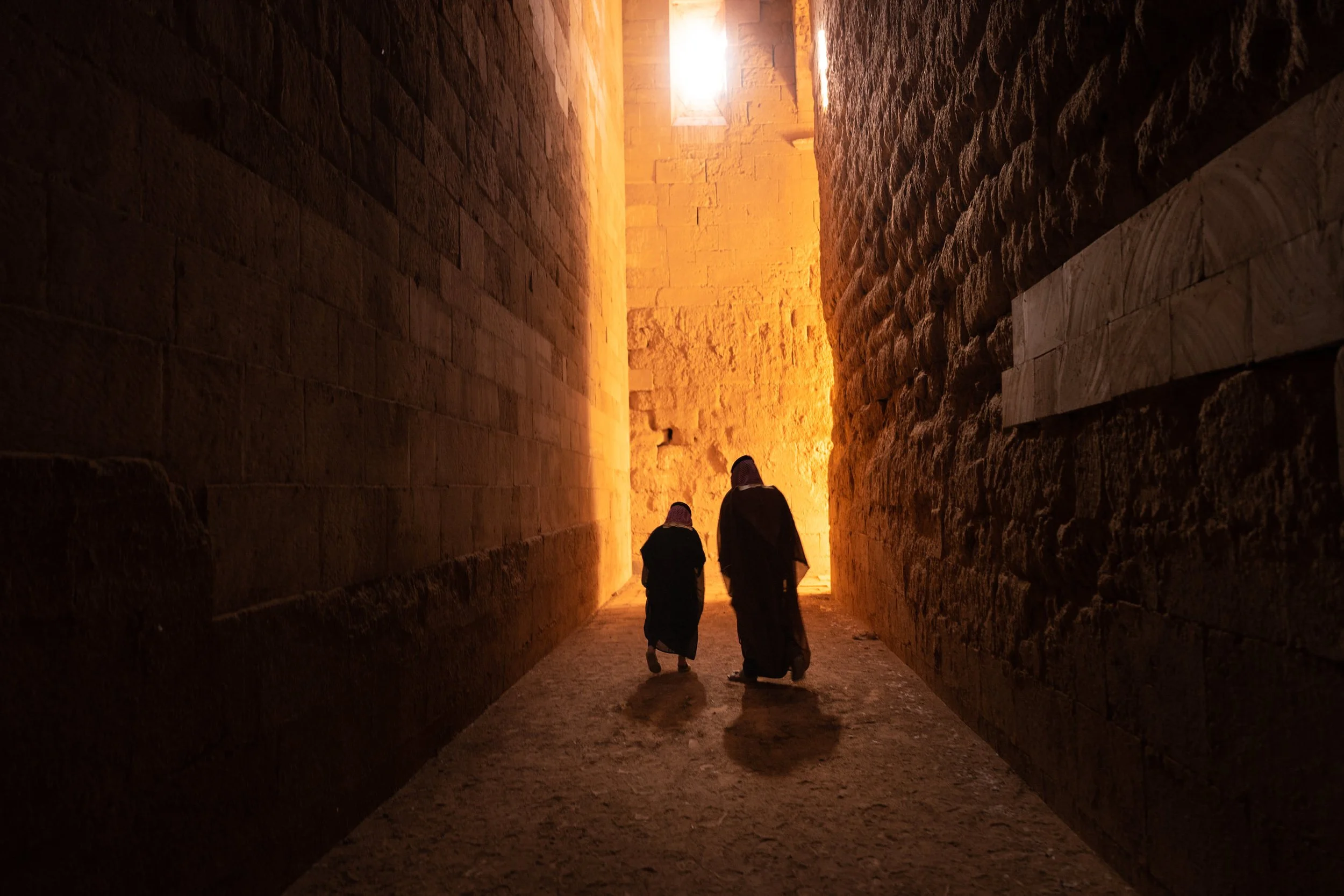 Two people walking through a narrow, ancient stone corridor with walls illuminated by warm sunlight from an opening above.