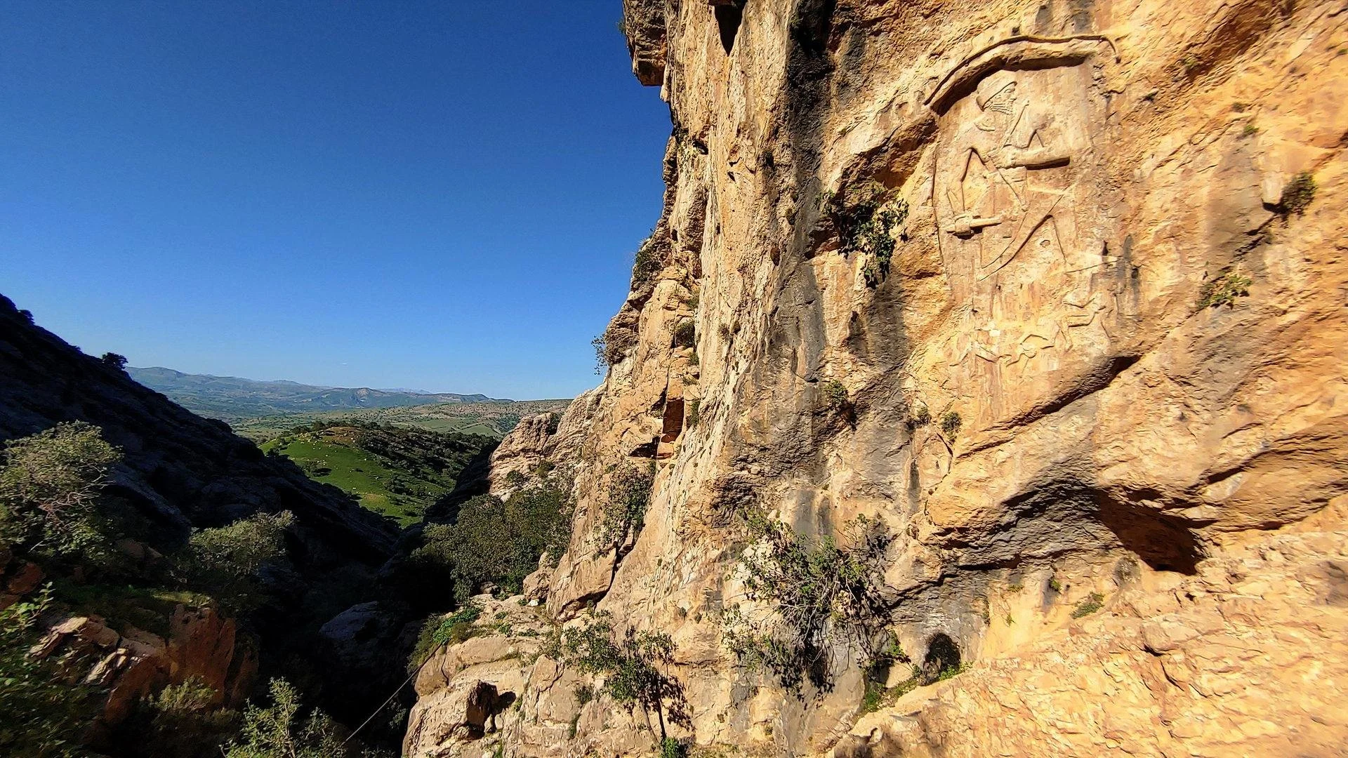Rocky cliff face with ancient carving of Bronze Age king under a clear blue sky.