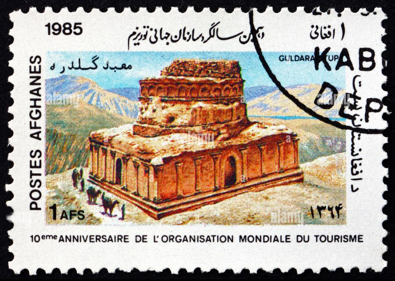 A postage stamp from Afghanistan issued in 1985, celebrating the 10th anniversary of the World Tourism Organization, featuring a drawing of a historical brick building with arches, set against a mountainous landscape.