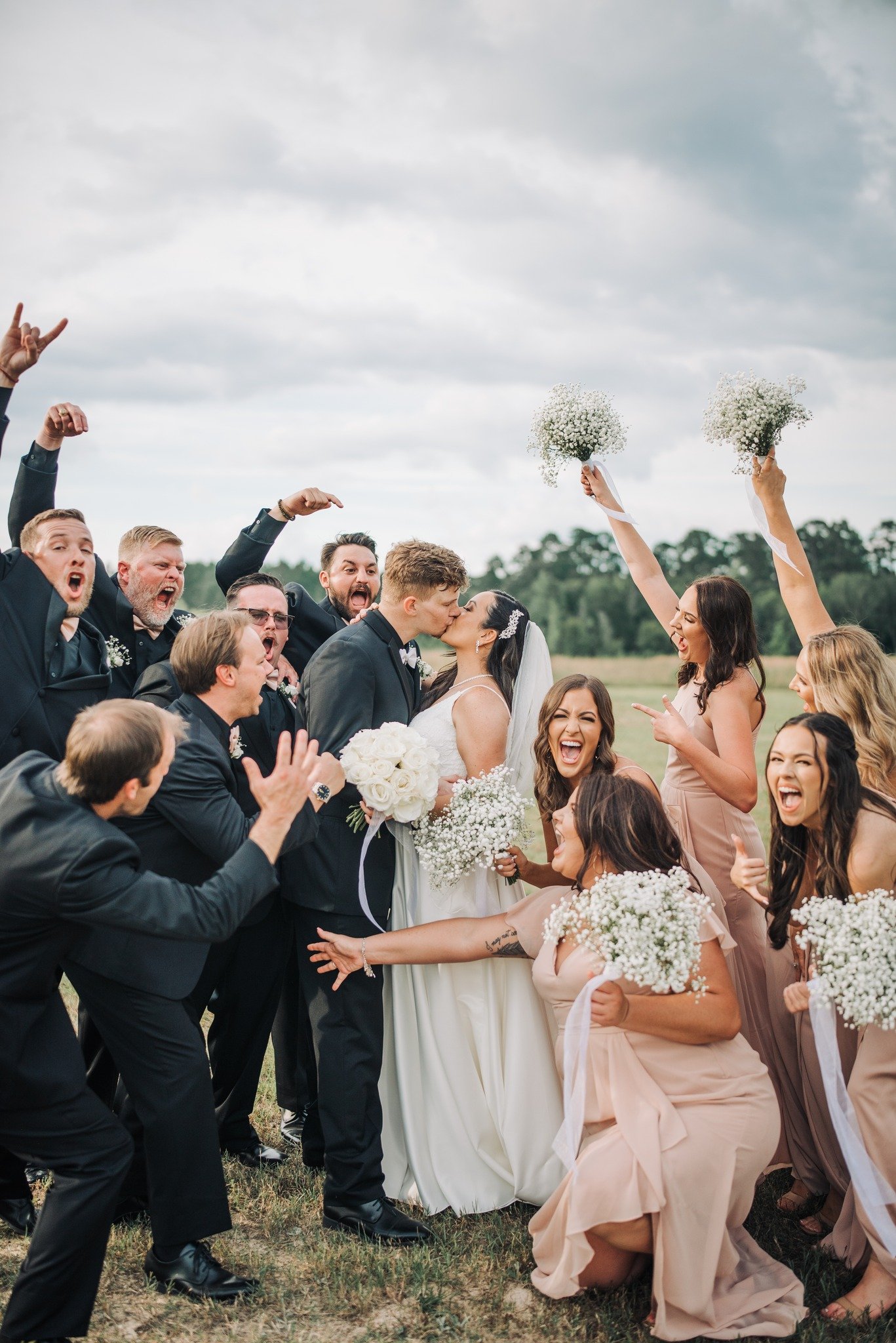 A wedding celebration with a bride and groom kissing, surrounded by happy wedding guests in an open field, some holding bouquets and others cheering.