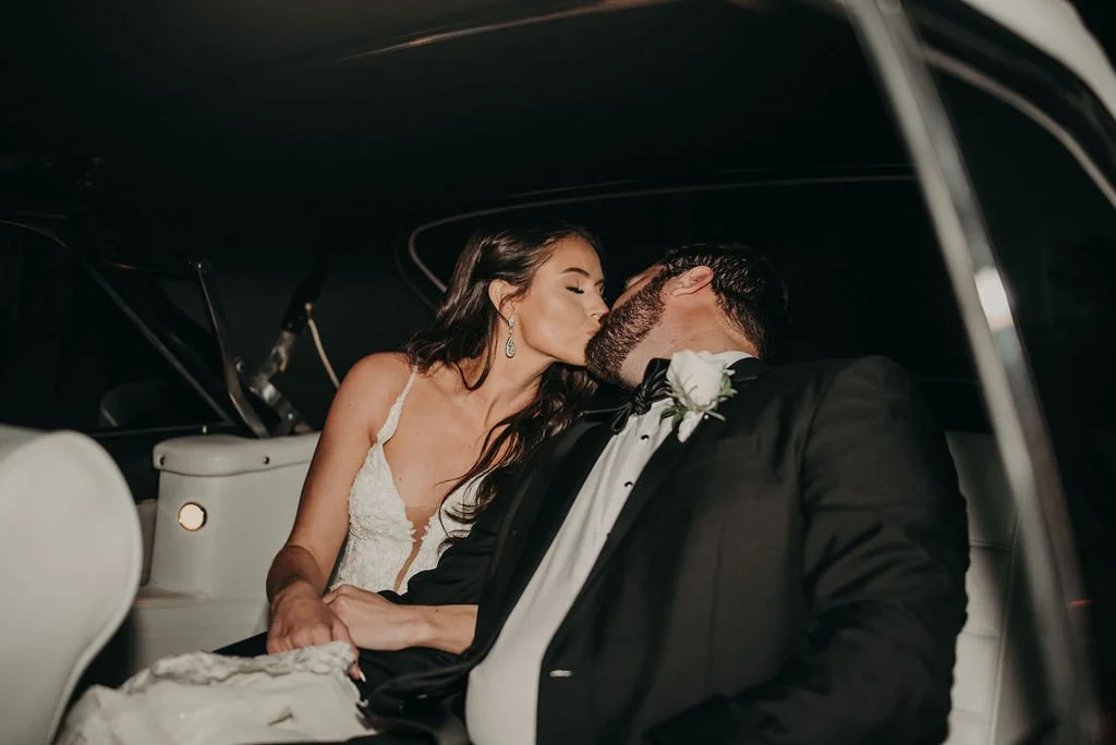A bride and groom sharing a kiss inside a car, with the bride in a wedding dress and the groom in a tuxedo with a white boutonnière.