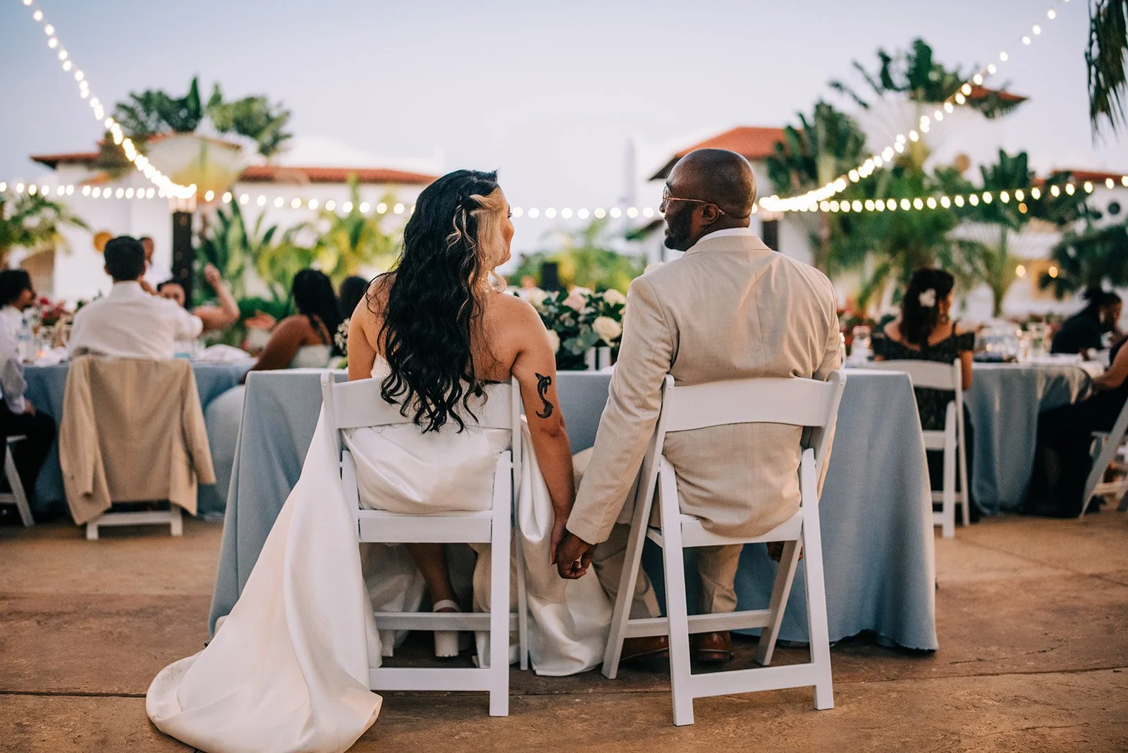 A newlywed couple sitting hand-in-hand at their wedding reception, with guests and string lights in the background, outdoors during evening. Punta Cana, Dominican republic wedding photographer