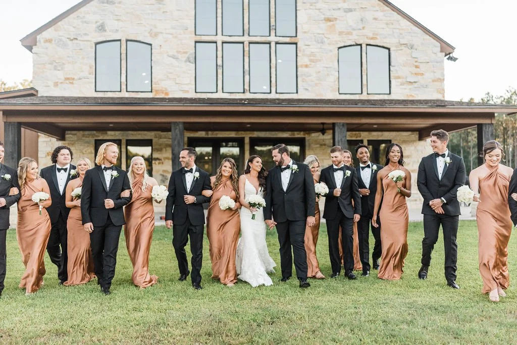 A wedding party walking outdoors in front of a stone building, with the bride and groom in the center, surrounded by bridesmaids in bronze gowns and groomsmen in black tuxedos, all smiling and holding flowers.