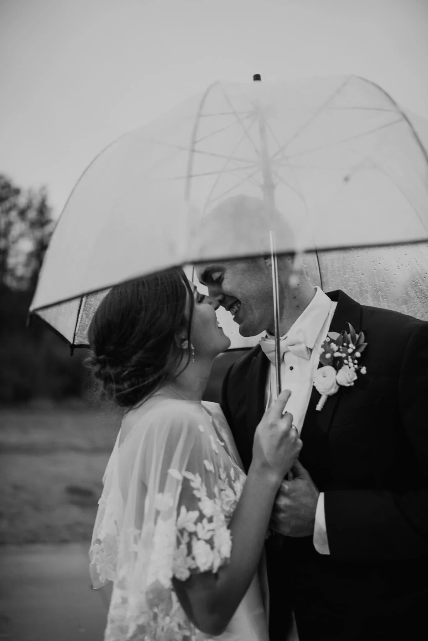 A black-and-white photo of a couple in wedding attire sharing an intimate moment under a transparent umbrella during rainy weather.
