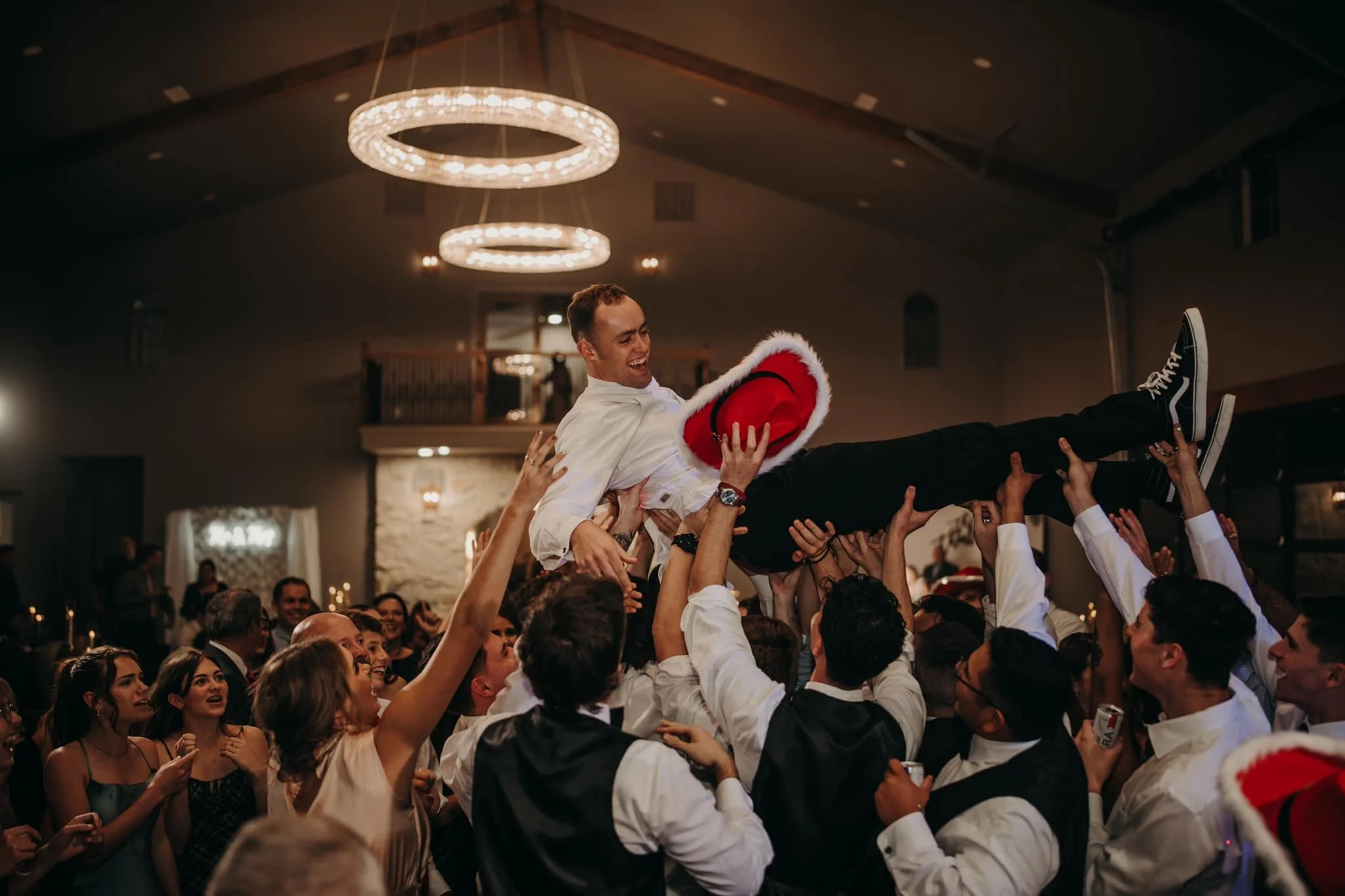A man wearing a white shirt and black pants is being crowdsurfed, holding a red Santa hat, at a celebration event with many smiling people around him, under warm lighting.