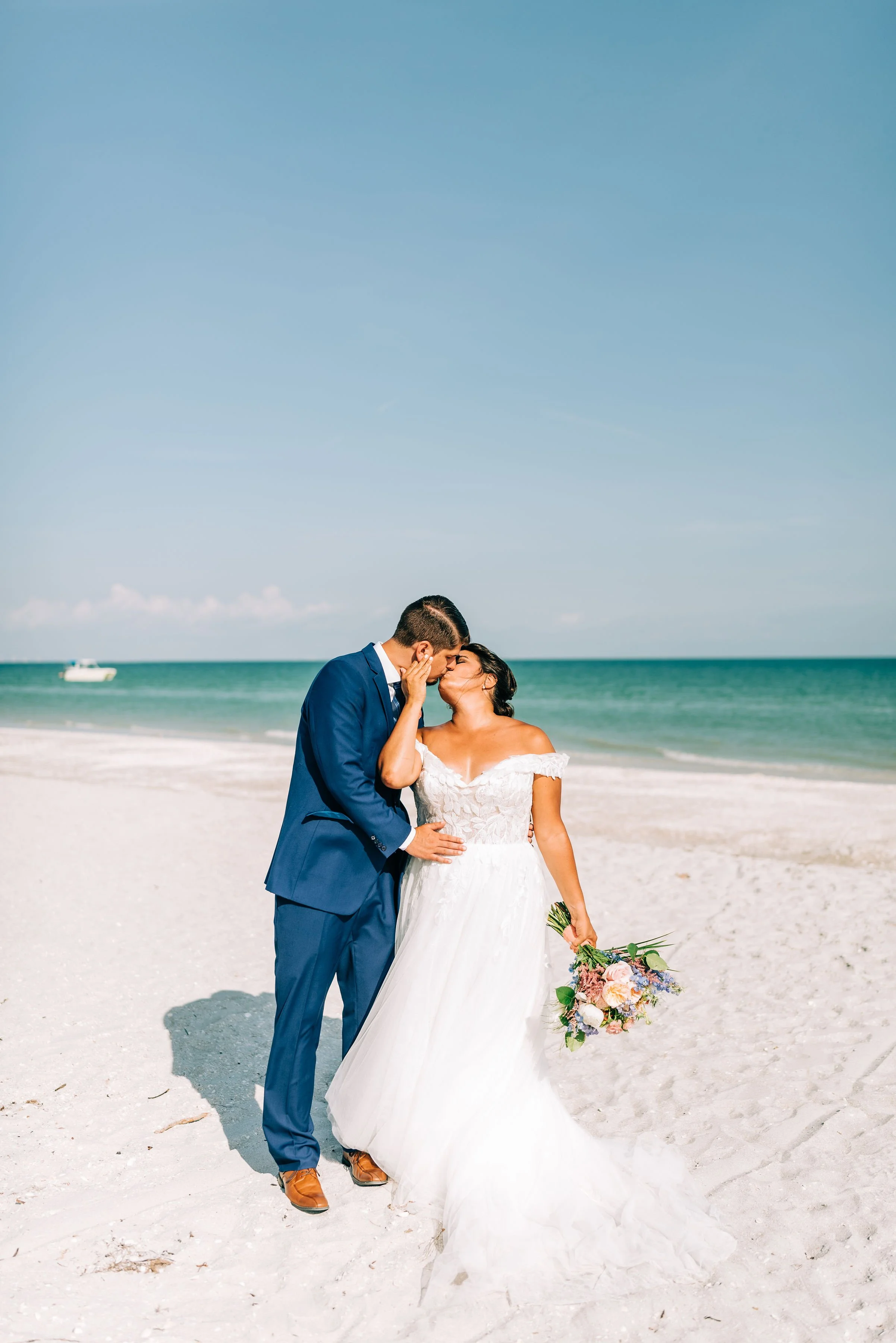 A bride and groom share a kiss on the beach during their wedding, with the ocean in the background and the bride holding a bouquet of flowers. Florida weddings