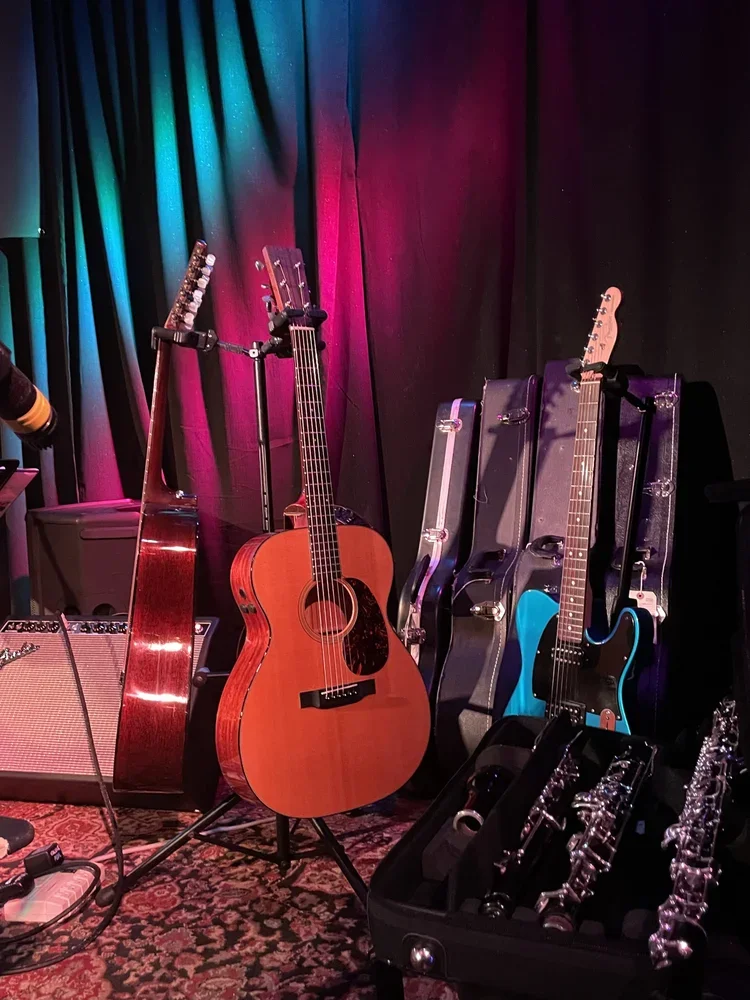 Acoustic and electric guitars with guitar cases, an amplifier, and clarinets on a patterned rug on stage with colorful curtain backdrop.