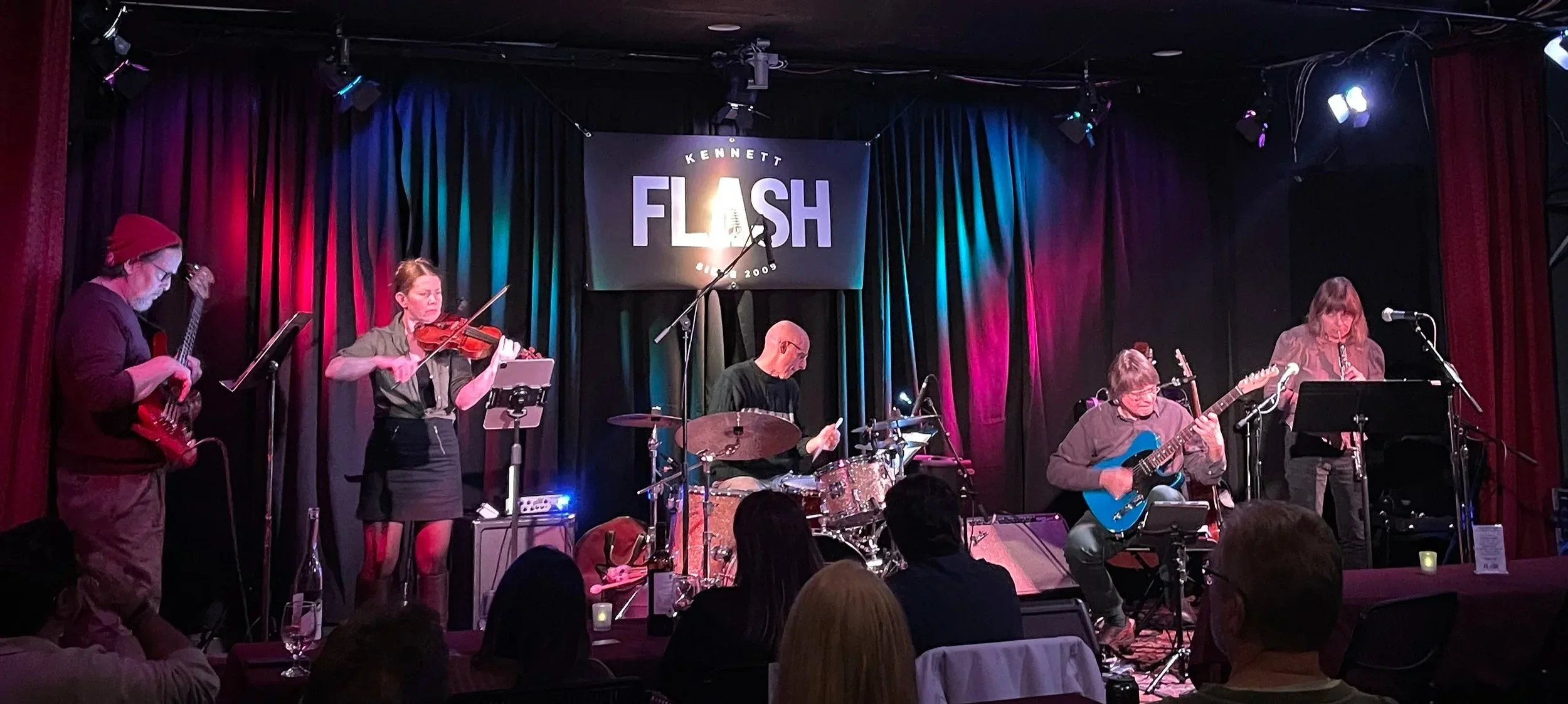 A band performing on stage at Kennedy Flash event in 2009, with musicians playing guitars, drums, a violin, and a flute, against colorful stage curtains and a sign overhead.