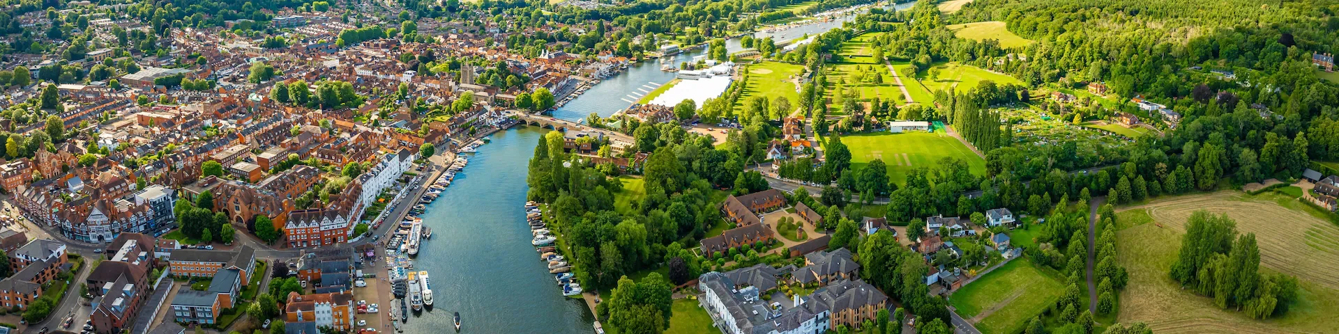 Aerial view of a town with a river running through it, with boats docked along the riverbank, surrounded by green parks, residential houses, and open fields, under a bright, sunny sky.