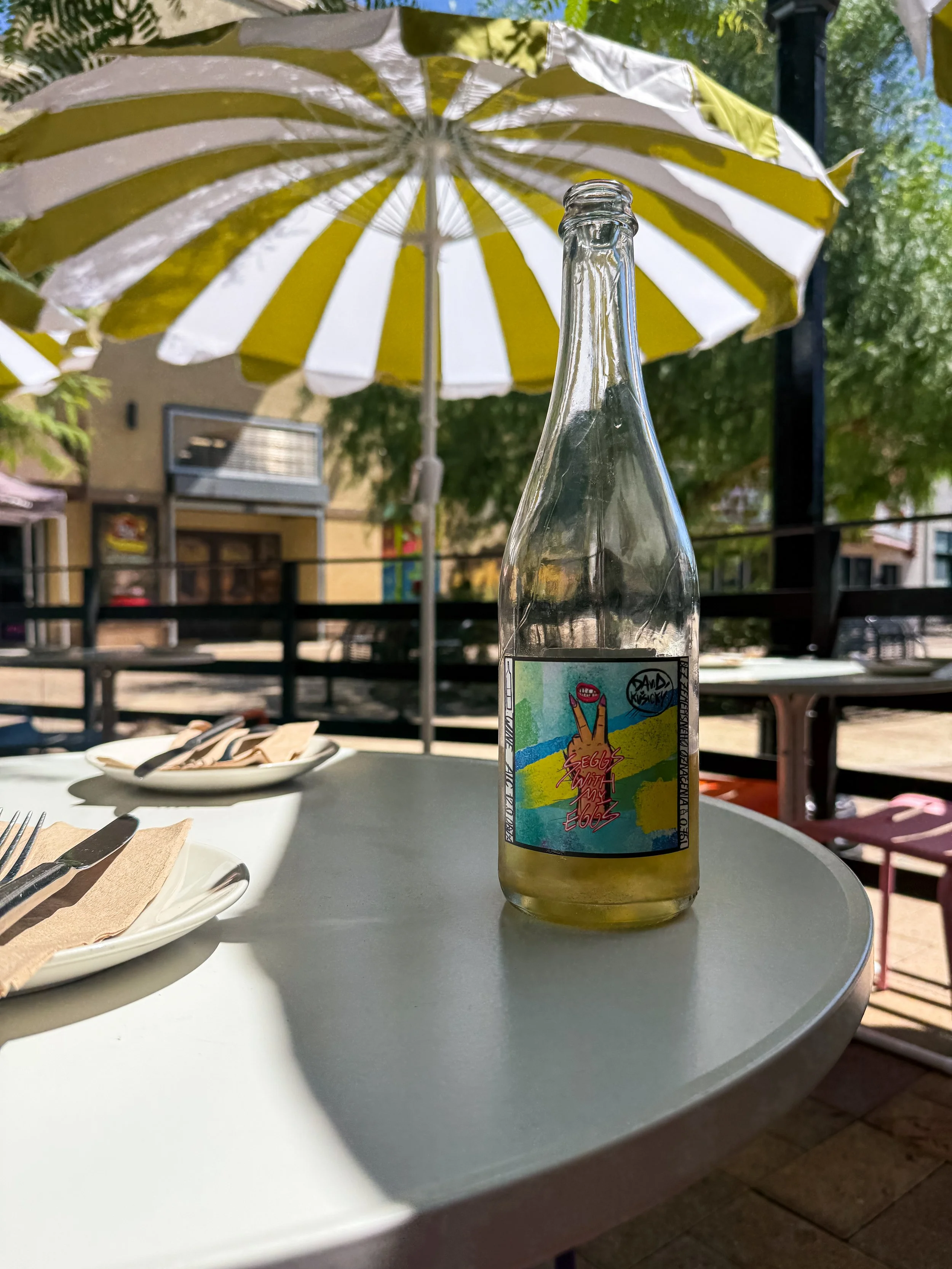 A clear glass soda bottle with a colorful label featuring an illustration of a hand making a peace sign, sitting on a white outdoor table. In the background, there is a yellow and white striped patio umbrella, some green trees, a building, and part o