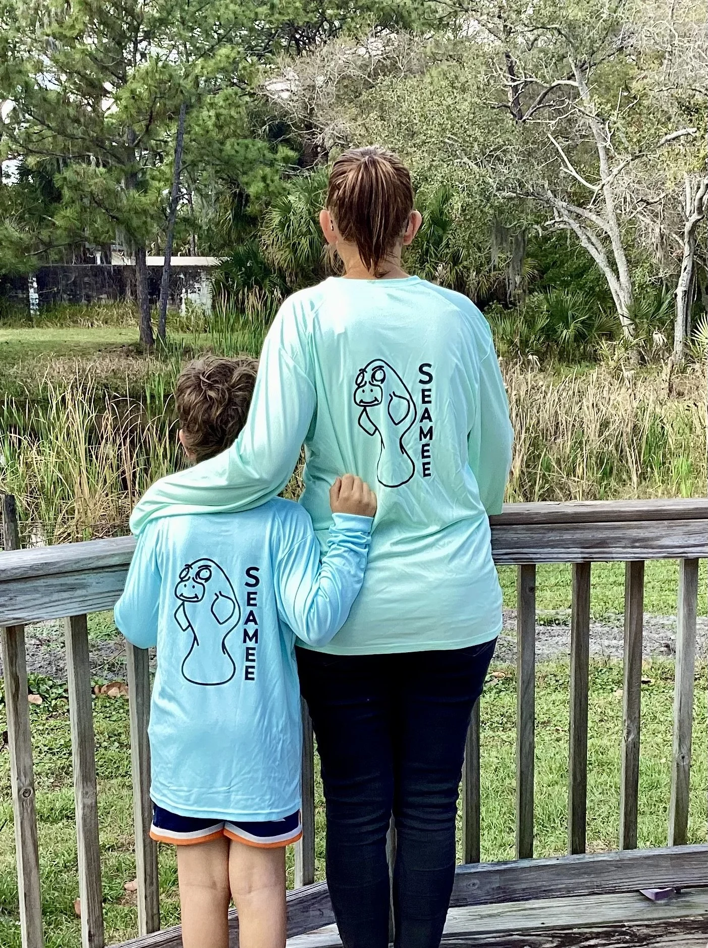 A woman and a child wearing matching teal shirts with a cartoon manatee and the word 'SEAMEE' printed on the back, standing on a wooden deck overlooking a grassy area with trees.