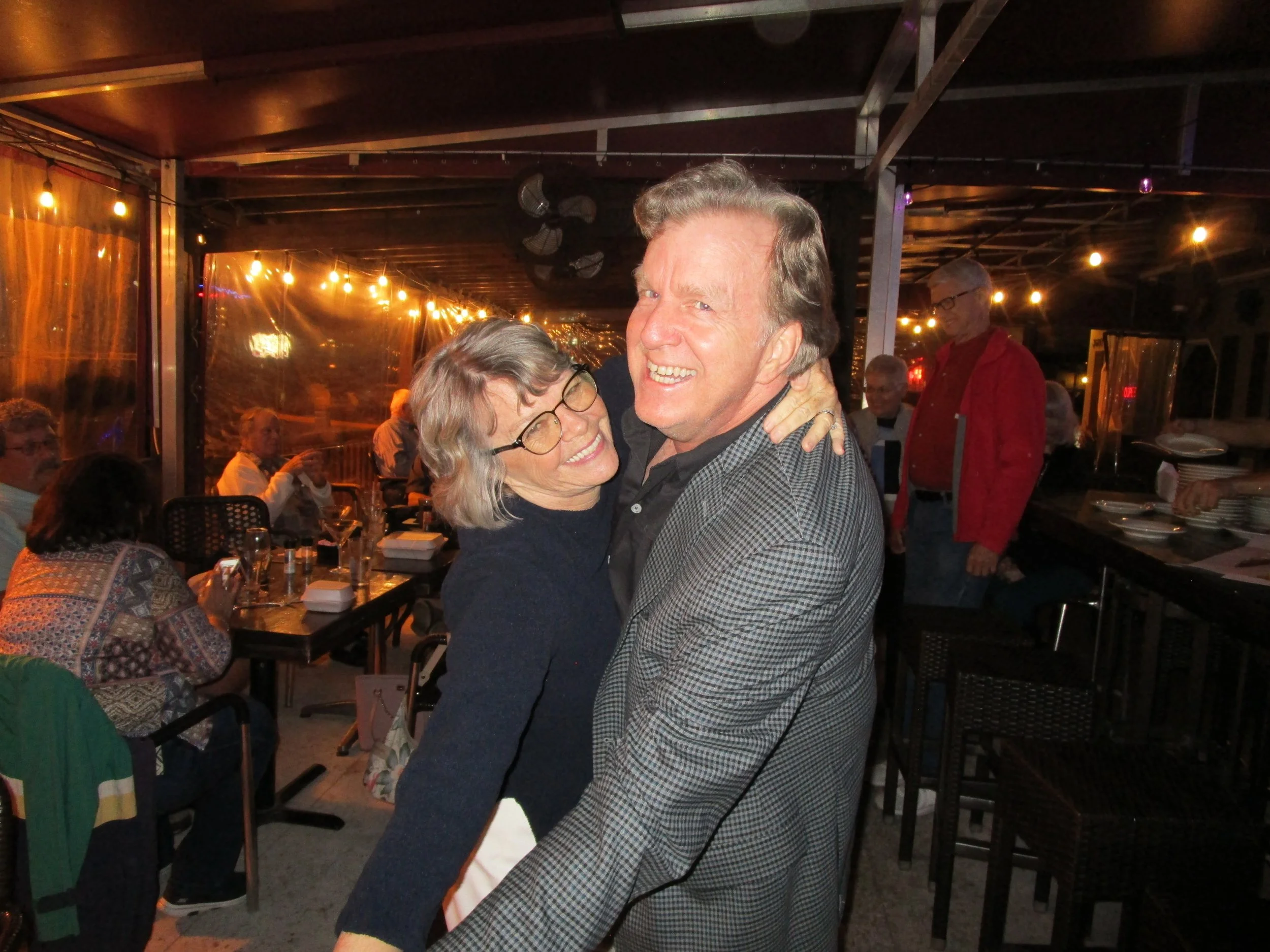 A man and woman smiling and dancing at a restaurant with string lights and other people dining in the background.