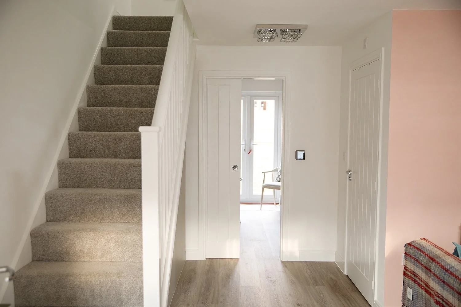 Interior of a house entryway with a carpeted staircase on the left, white walls, a glass door at the back, and a pink wall on the right side. There is a small chair visible through the glass door and a sofa with a colorful blanket on the right.