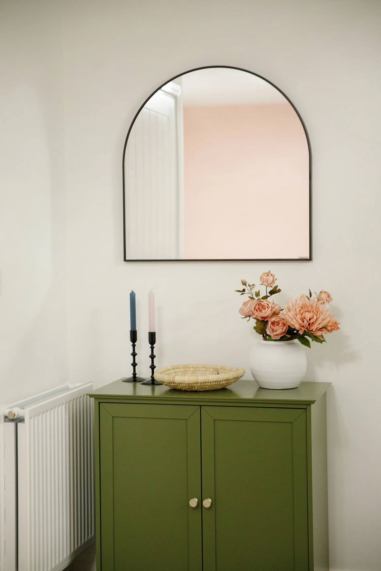 A green cabinet with a vase of pink flowers on top, flanked by two candle holders with pink and blue candles, a woven basket, a white radiator and a mirror on the wall