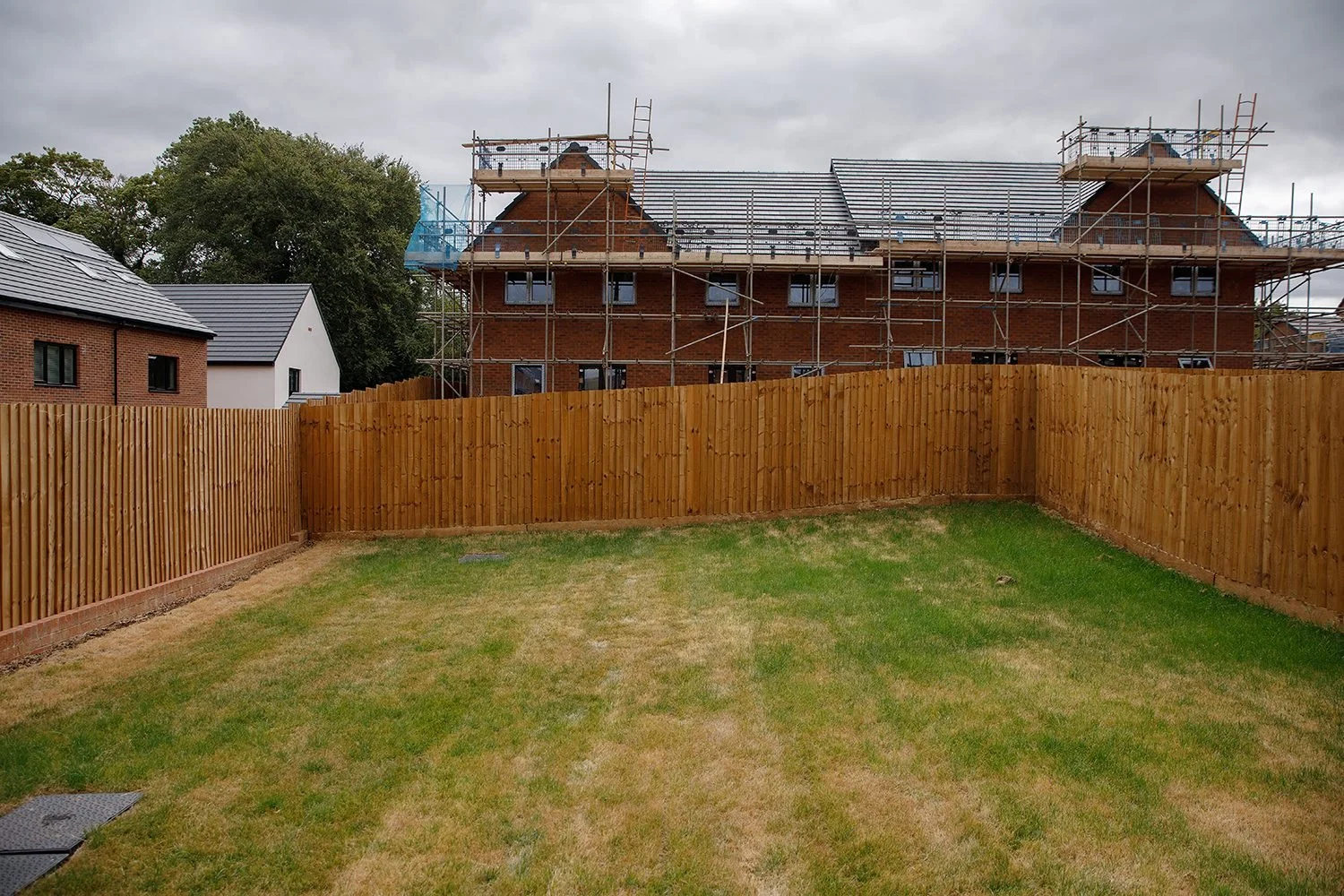 View of a backyard with newly installed wooden fence, adjacent to a construction site with a brick building under renovation and scaffolding on a cloudy day.