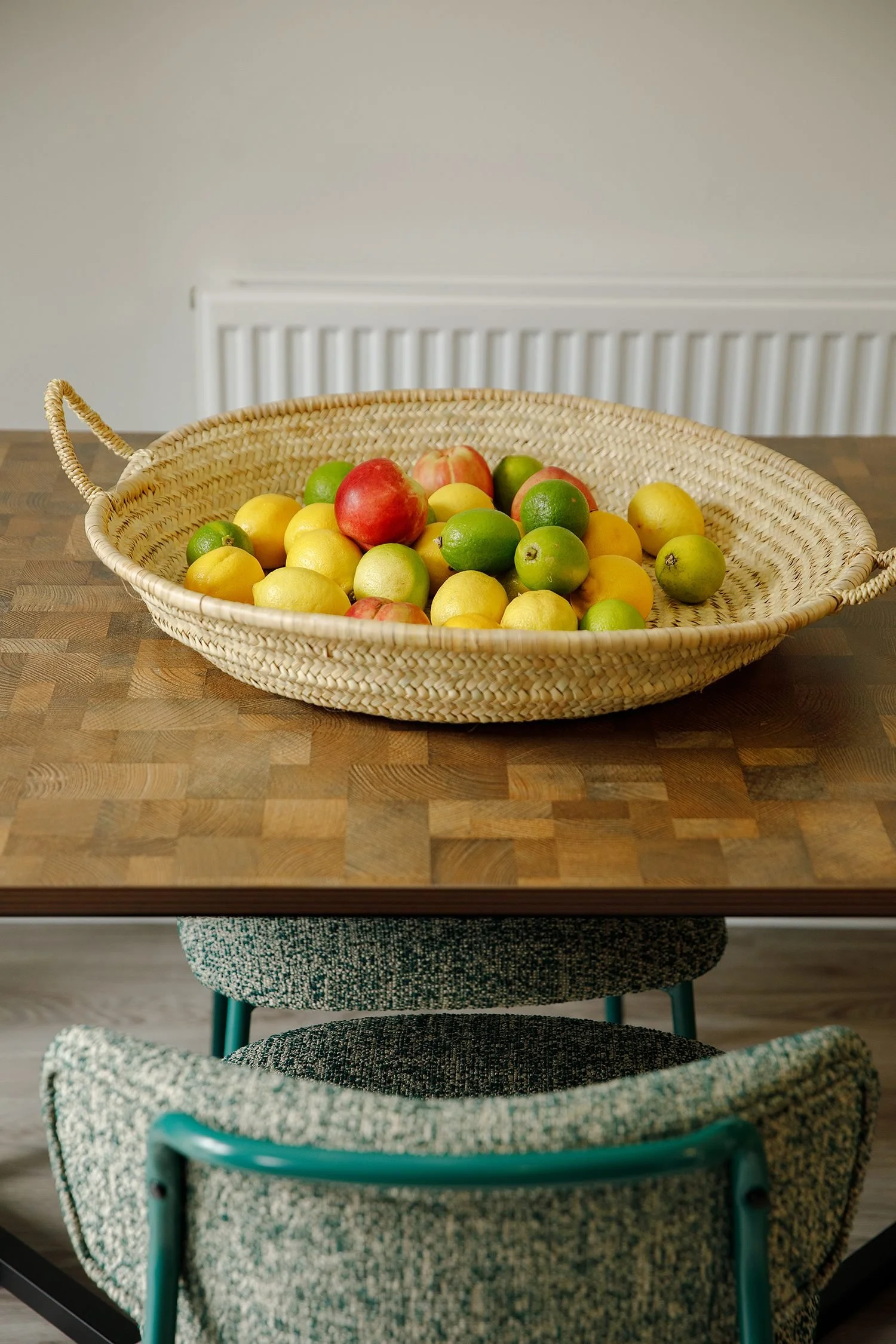 A wicker basket filled with apples, lemons, and limes is on a wooden table. A radiator is visible on the beige wall behind the basket. The table is surrounded by a patterned chair.