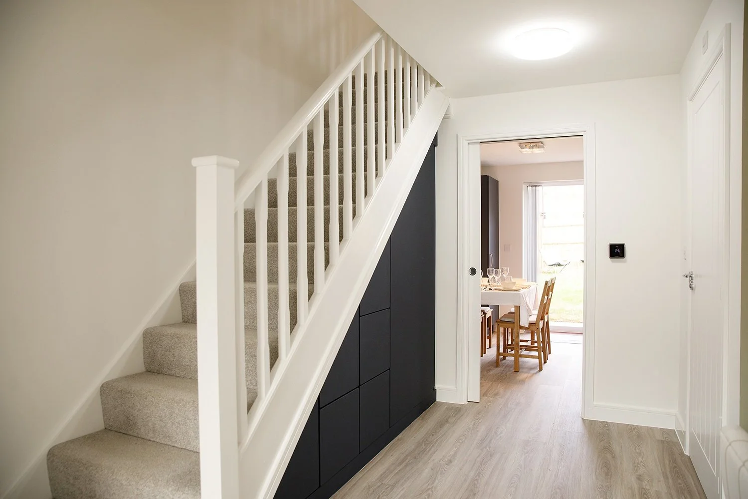 Interior of a modern home with a carpeted staircase, black storage cabinets beneath the stairs, and a doorway leading to a dining area with a table set for a meal and sliding glass door to the outdoors.