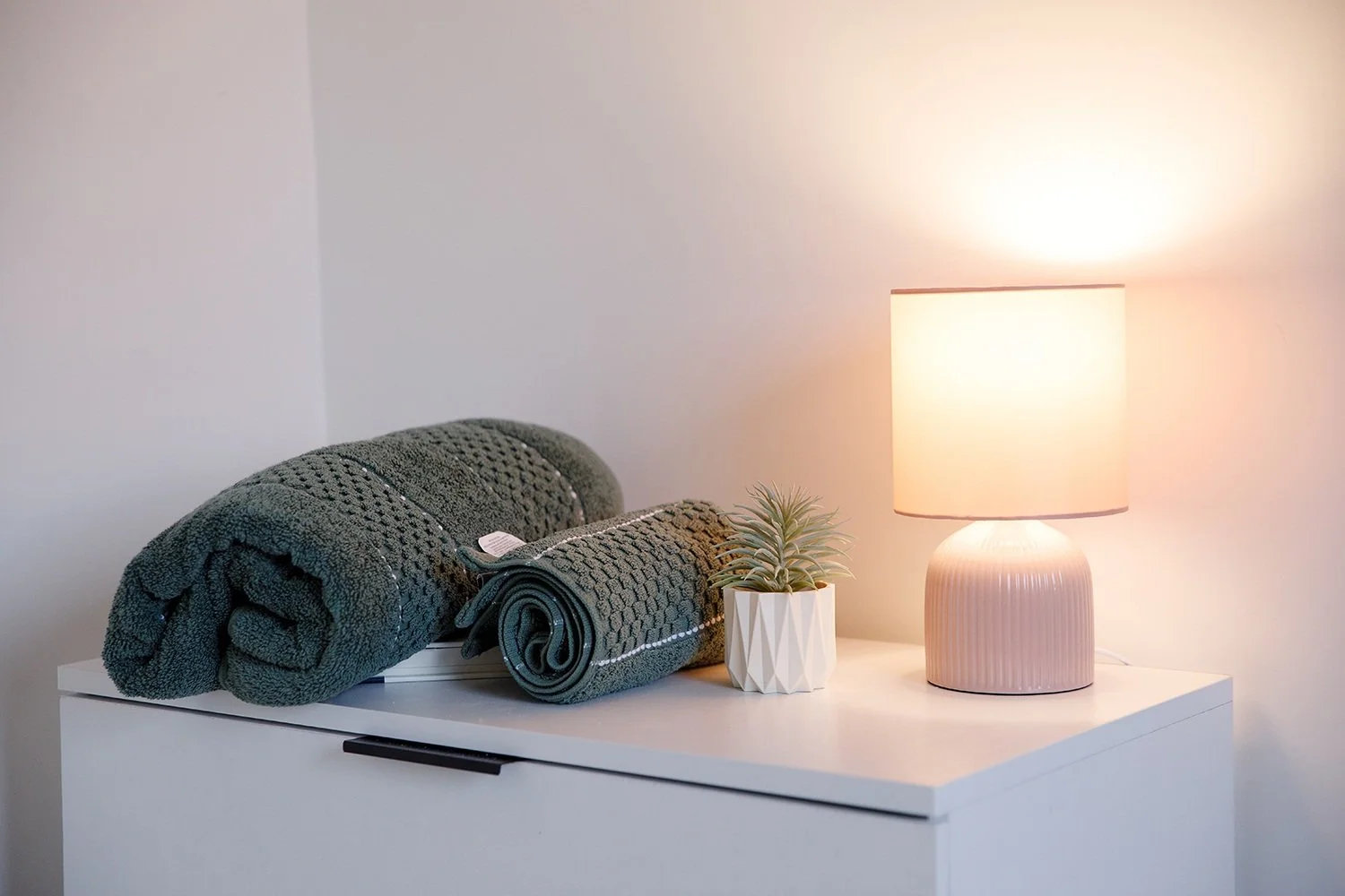 A white dresser with two rolled green textured towels, a small potted succulent, and a beige table lamp with a softly glowing shade against a plain wall.