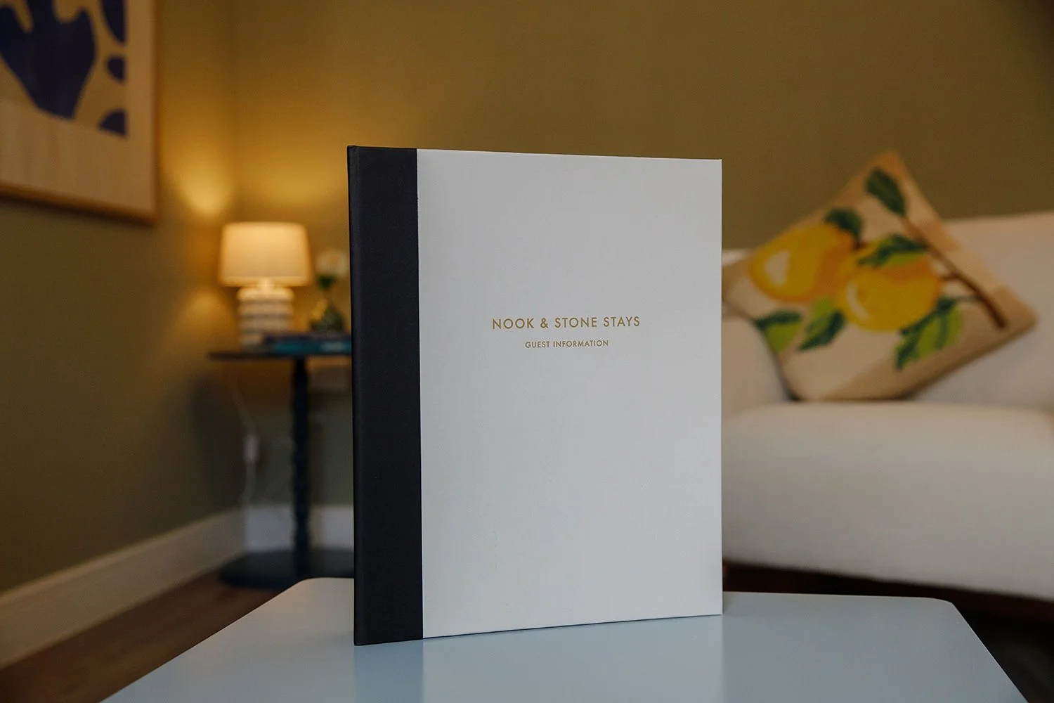 Guest information booklet titled "Nook & Stone Stays" placed on a white table in a cozy living room with a sofa, decorative pillow, and a side table with a lamp in the background.