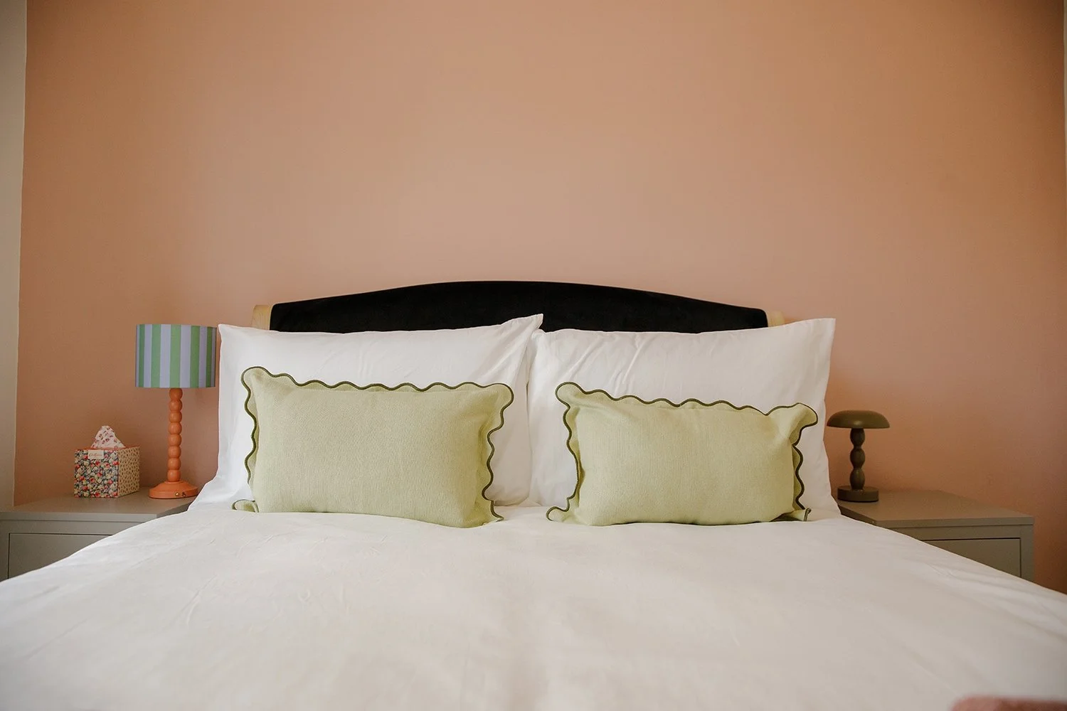 A neatly made bed with two white pillows, two green decorative pillows, a black headboard, pink wall, and bedside tables with lamps and a tissue box.