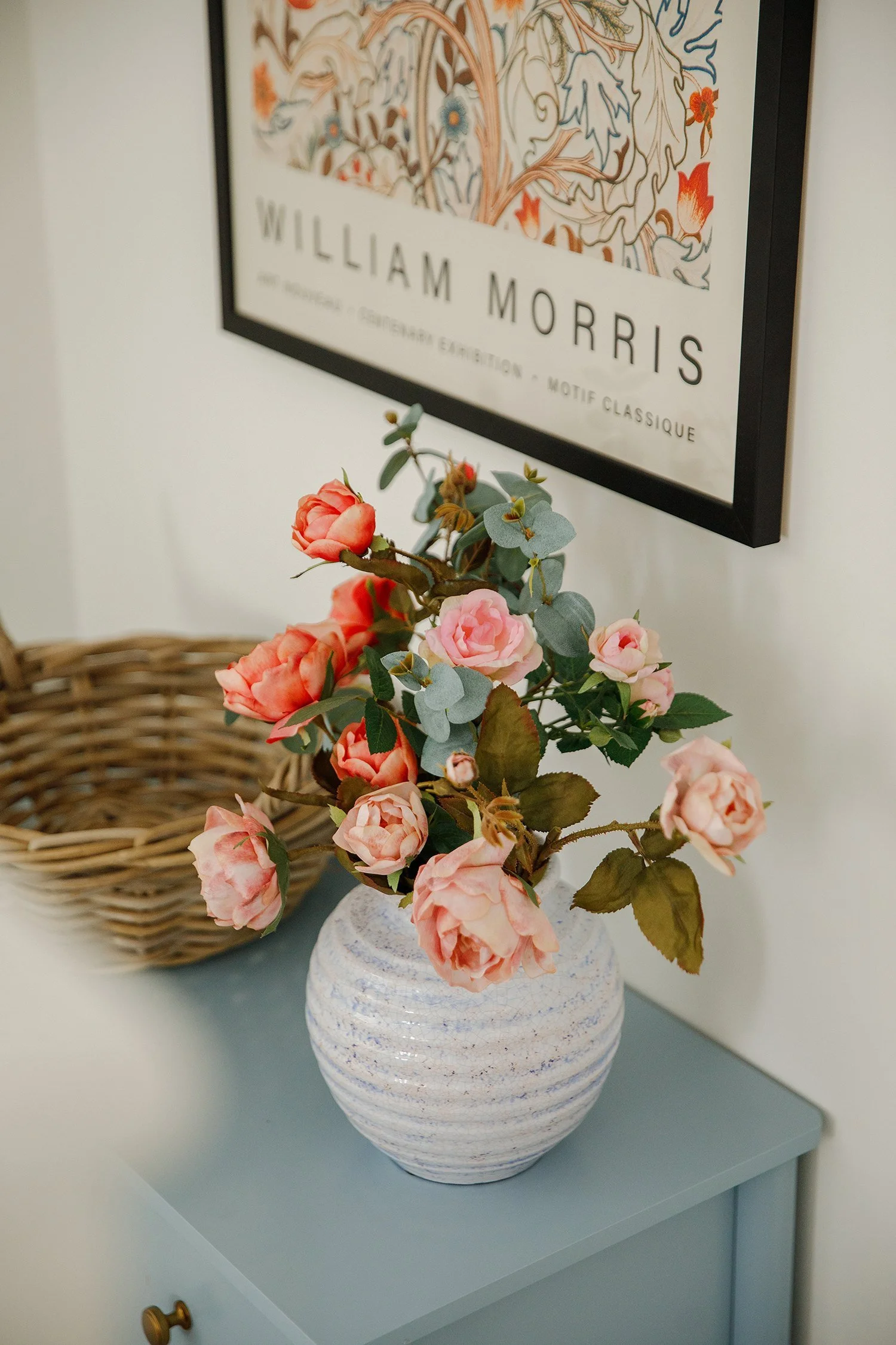 A white textured ceramic vase with pinkish-peach roses and eucalyptus leaves on a light blue surface. Behind it, a wicker basket and a framed William Morris print on a white wall.