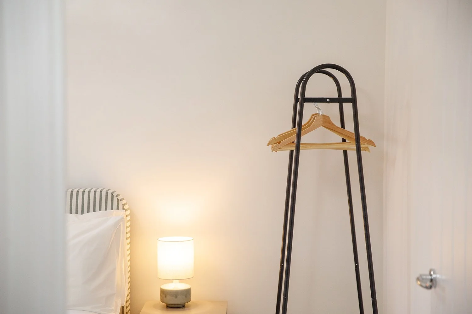 Part of a bedroom with a bedside table, a small lamp, and a black metal clothing rack with wooden hangers.