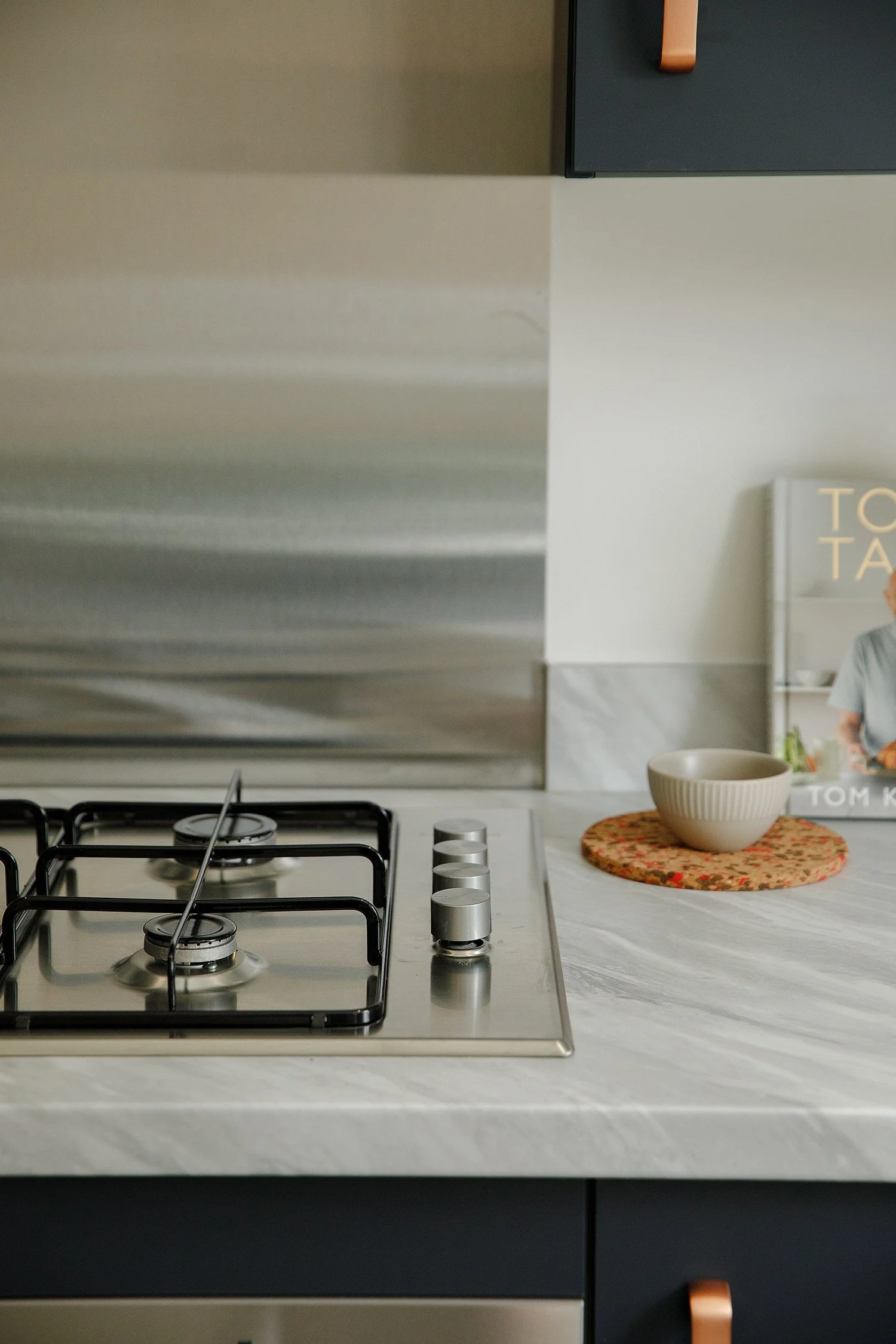 Kitchen with stovetop and a white marble countertop, with a beige bowl on a round floral placemat and a cookbook with a person on the cover.