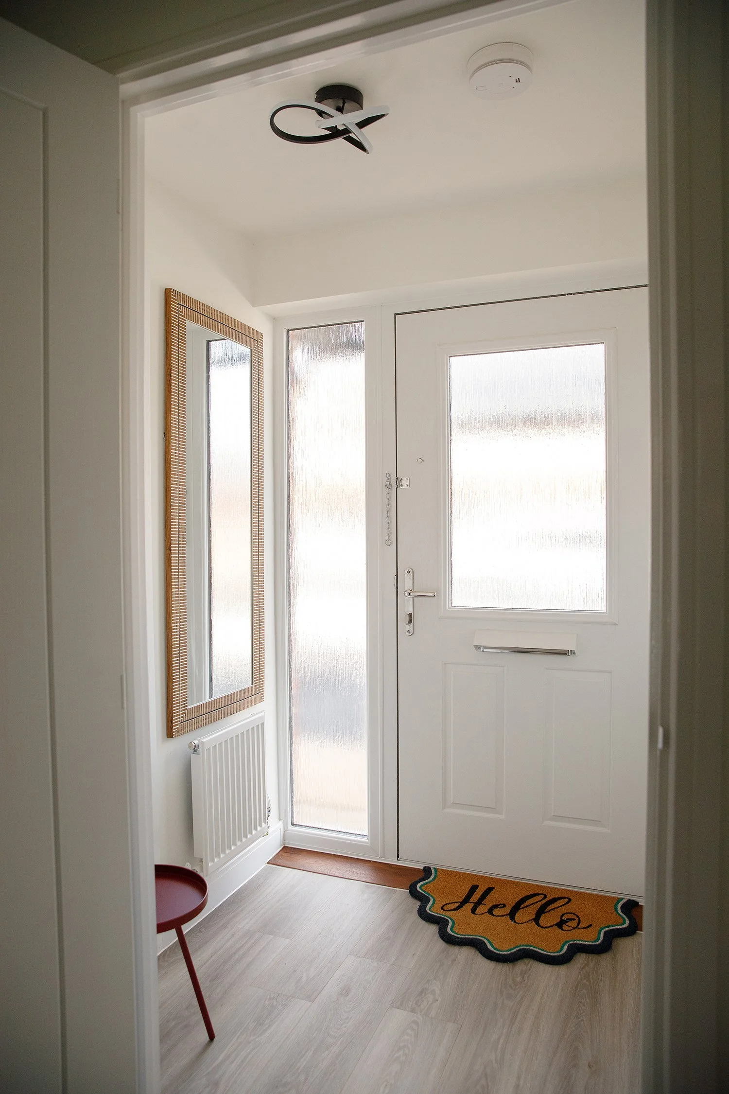 A front door with frosted glass panels, a 'Hello' welcome mat, a small red table, a wall mirror with a wooden frame, a radiator, and a hanging black modern ceiling light fixture.