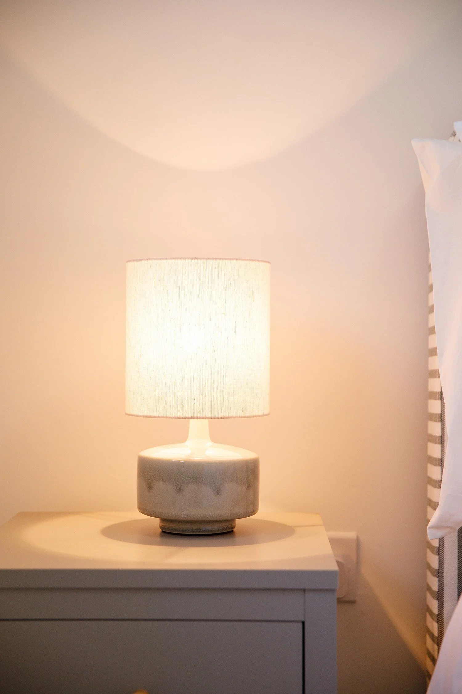 A table lamp with a cylindrical lampshade on a nightstand in a warmly lit room.