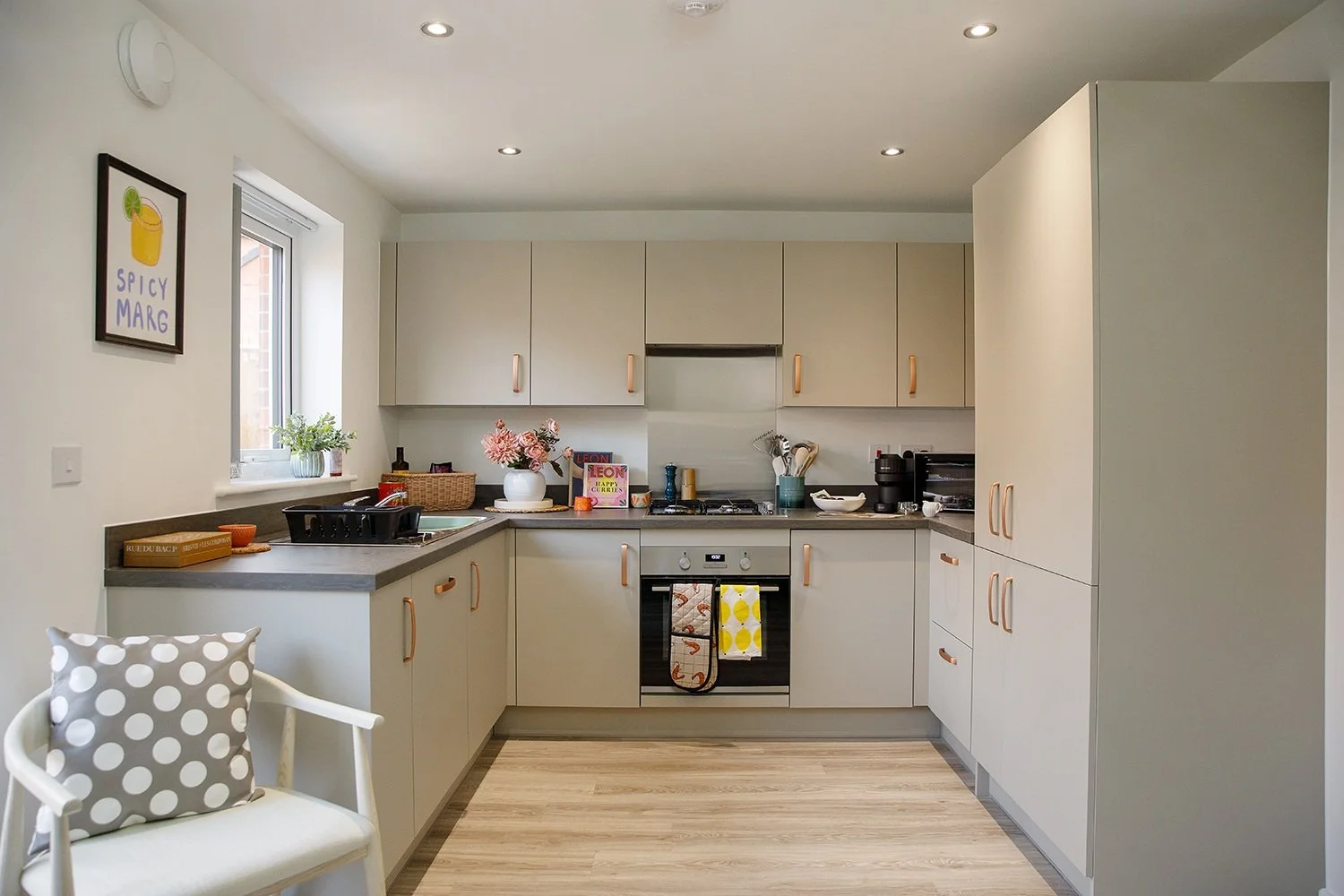 Modern kitchen with beige cabinets, black countertops, and a window with a plant. Decor includes pink flowers, a framed print of a lemon drink, and various kitchen essentials.