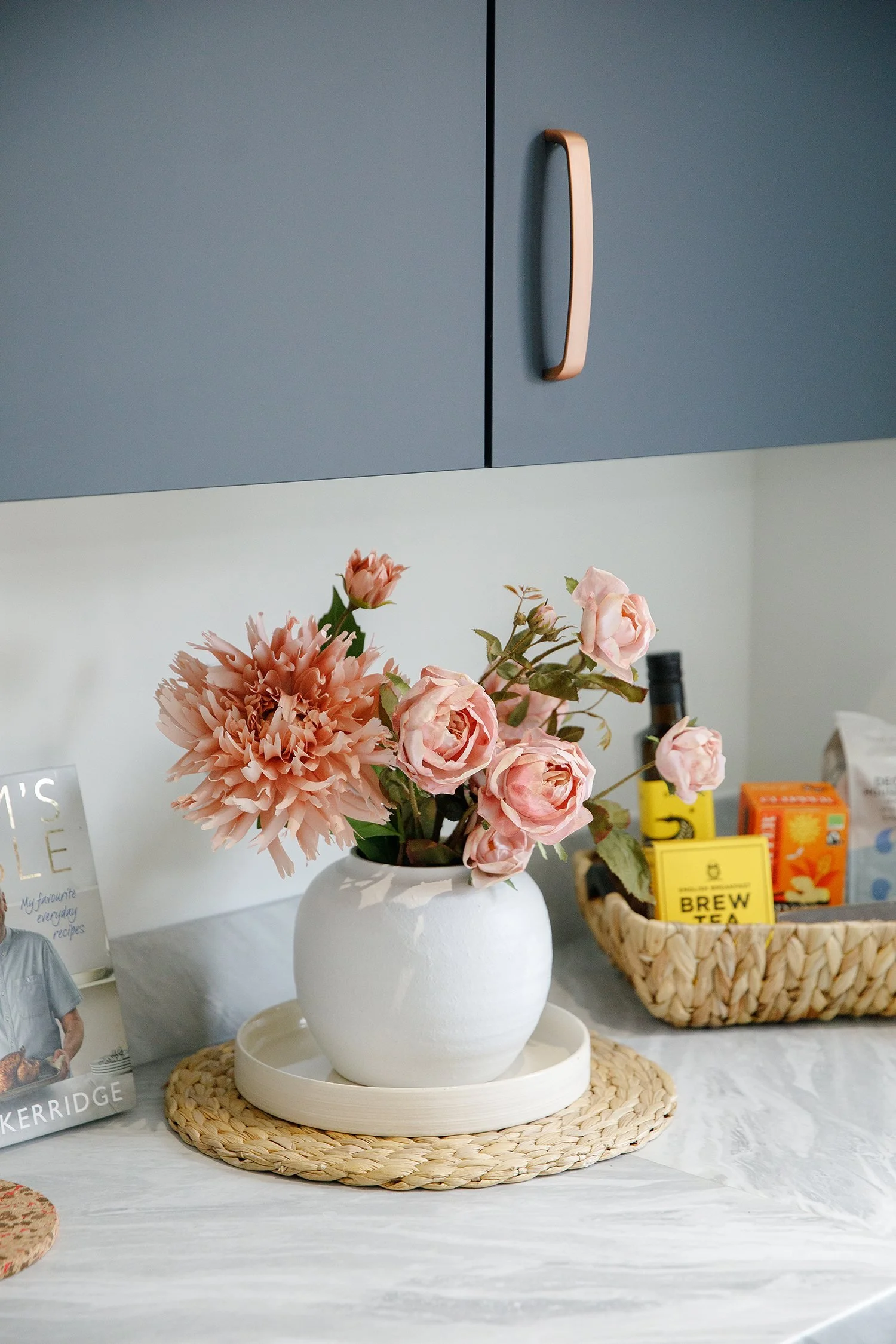 A white ceramic vase with pink roses and peony flowers on a woven straw placemat. The background includes tea boxes and a lattice basket with yellow and orange packaging.